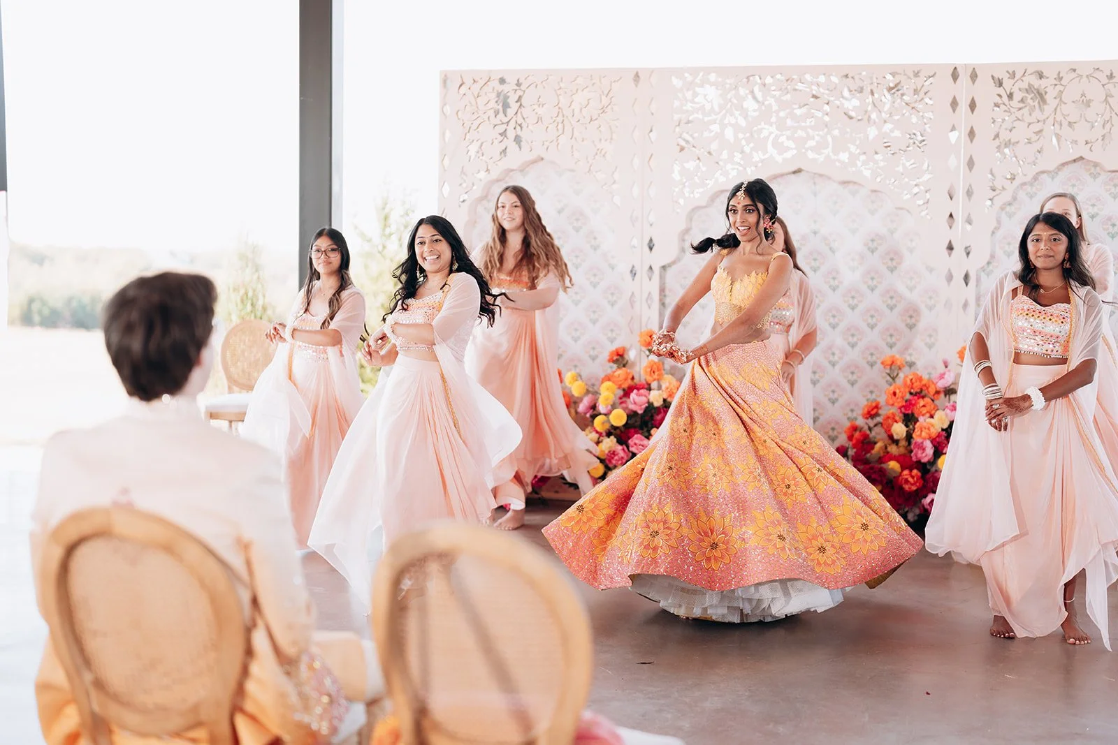 An Indian woman wearing a colorful, traditional Indian dress is dancing at a celebration, surrounded by women in pastel-colored dresses, with a floral backdrop and seating area in the background.