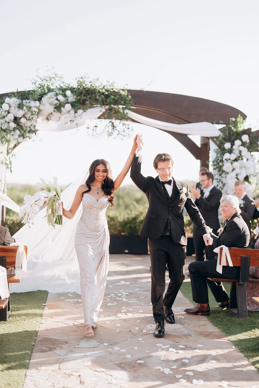 A newlywed Indian couple walking down the aisle together outdoors, holding hands with their arms raised, with guests clapping in the background and floral decorations overhead.