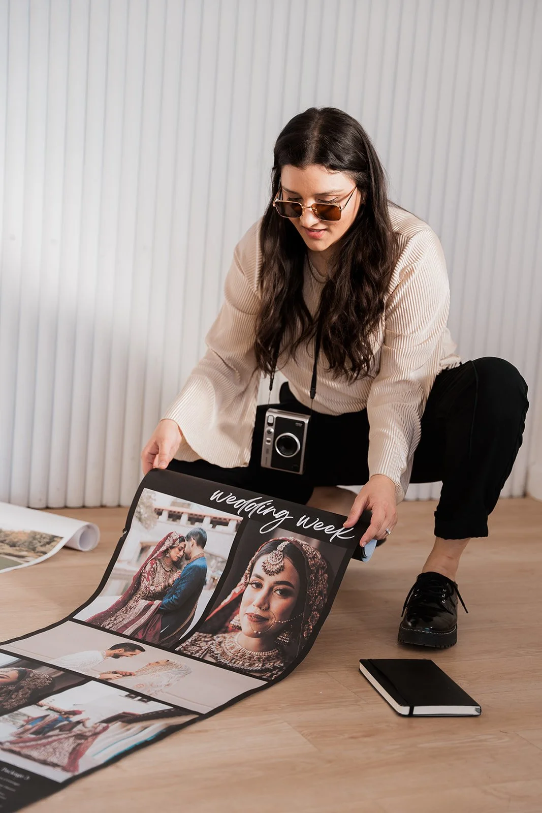 Woman with long dark hair, glasses, wearing a beige sweater, crouching on a wooden floor, holds a wedding photo album open showing pictures of a bride and groom, with a black notebook on the floor nearby.