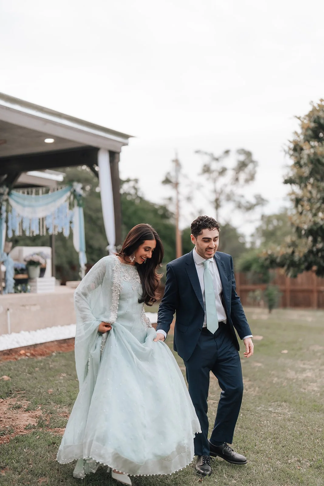 Pakistani Couple, bride in a white dress and groom in a navy suit, holding hands and walking on a grassy outdoor area during their wedding celebration.