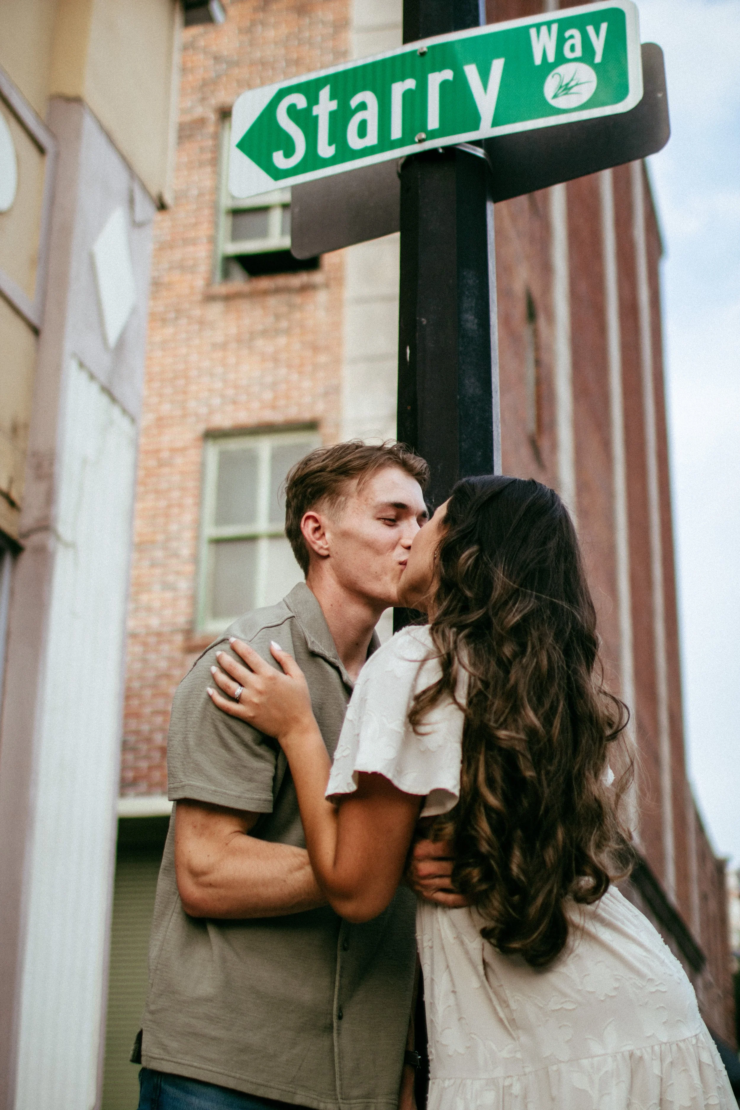 A couple sharing a kiss under a street sign that says 'Starry Way' in an urban setting.