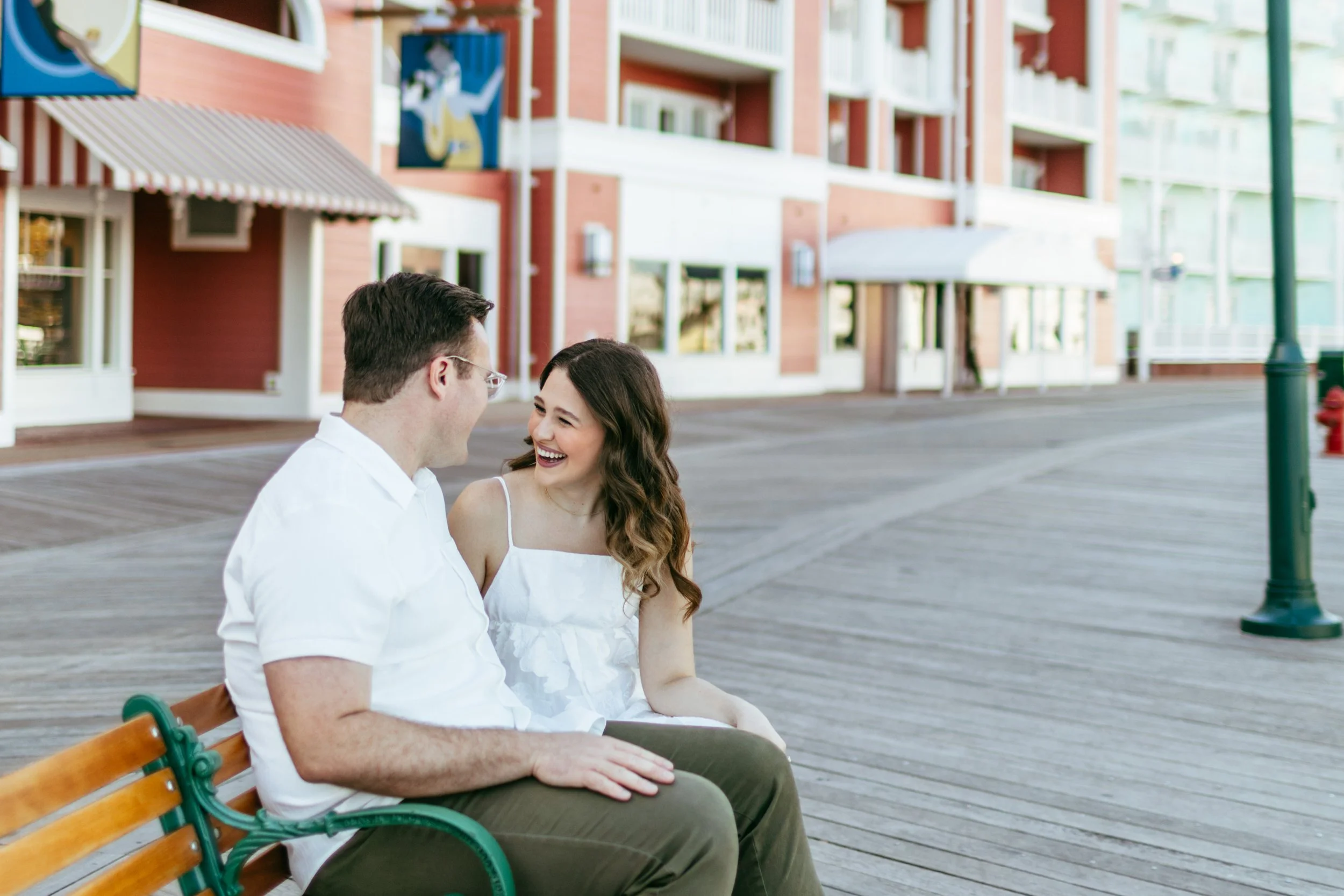 A man and woman sitting on a park bench in a shopping or residential area, smiling and talking to each other with red brick buildings and shops in the background.