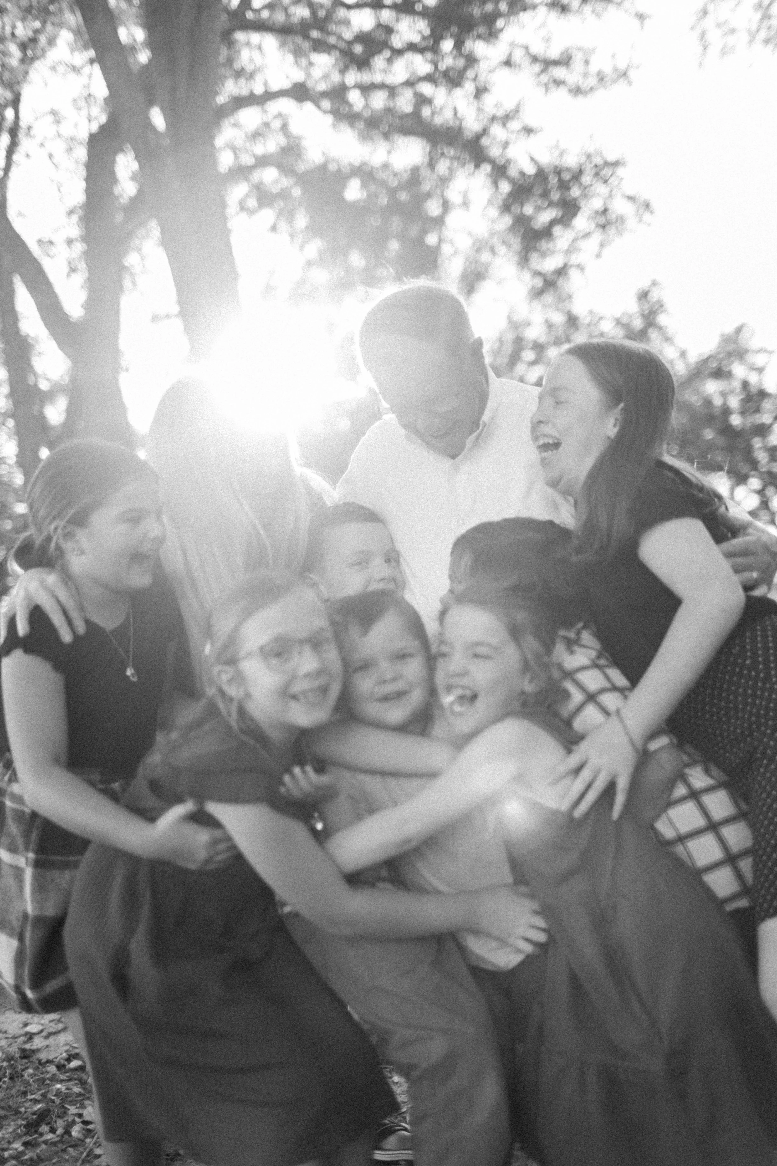 A group of children and two adults hugging and smiling outdoors with sunlight shining through trees in the background.