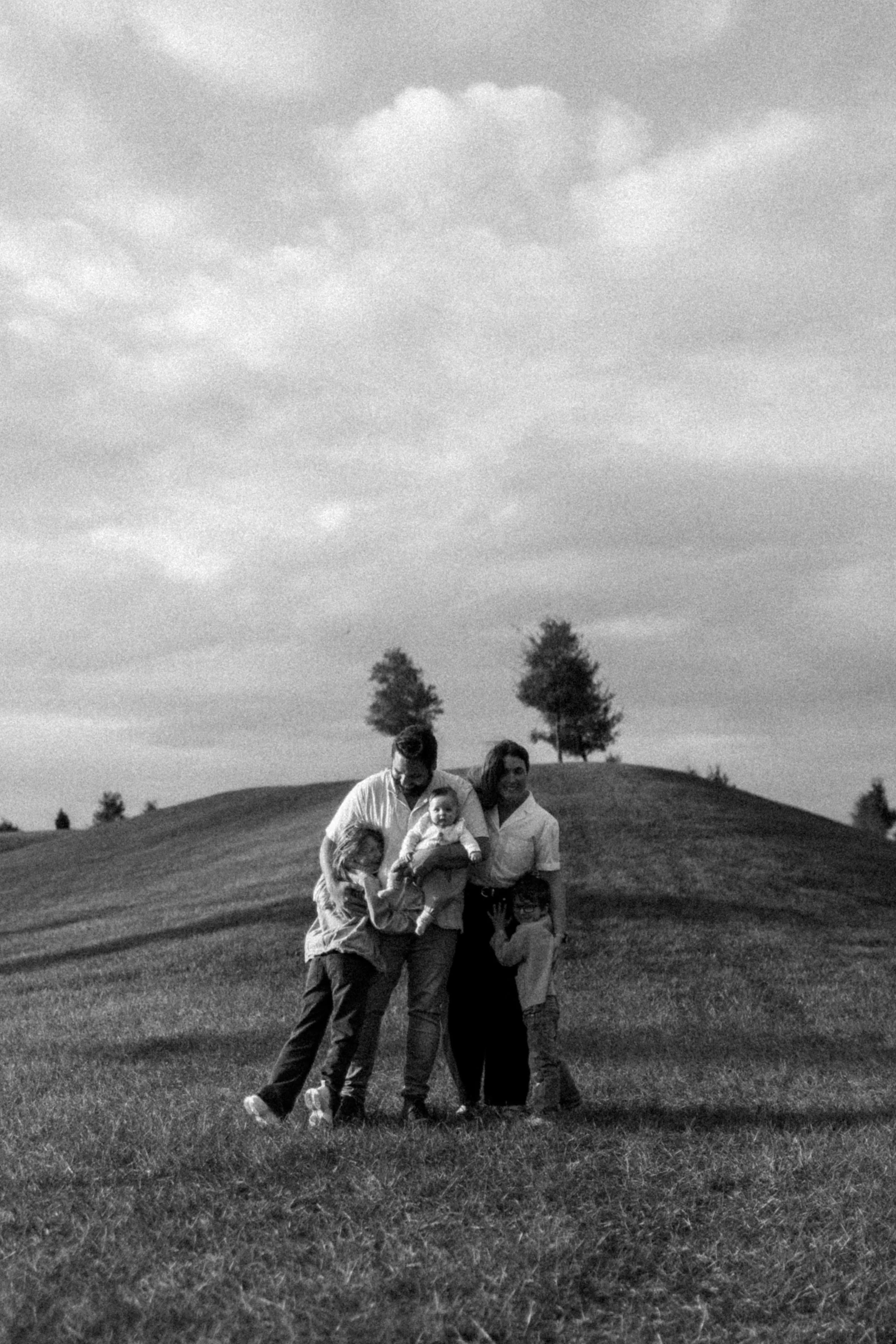 A black and white photo of a family of five standing on a grassy hill with two trees in the background and a cloudy sky.
