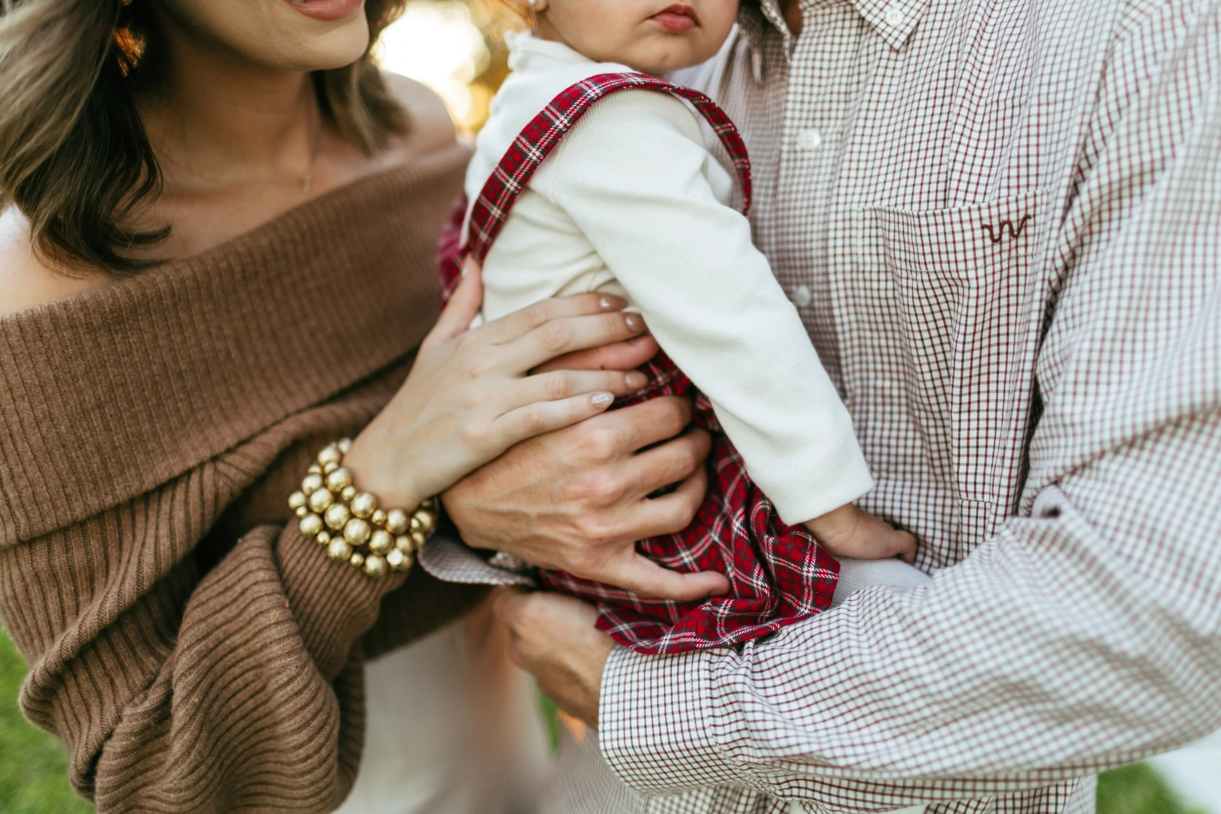 A woman with a brown off-shoulder top wearing gold pearl bracelets holding a young girl in a red plaid dress while a man in a checkered shirt supports the girl with his hands.