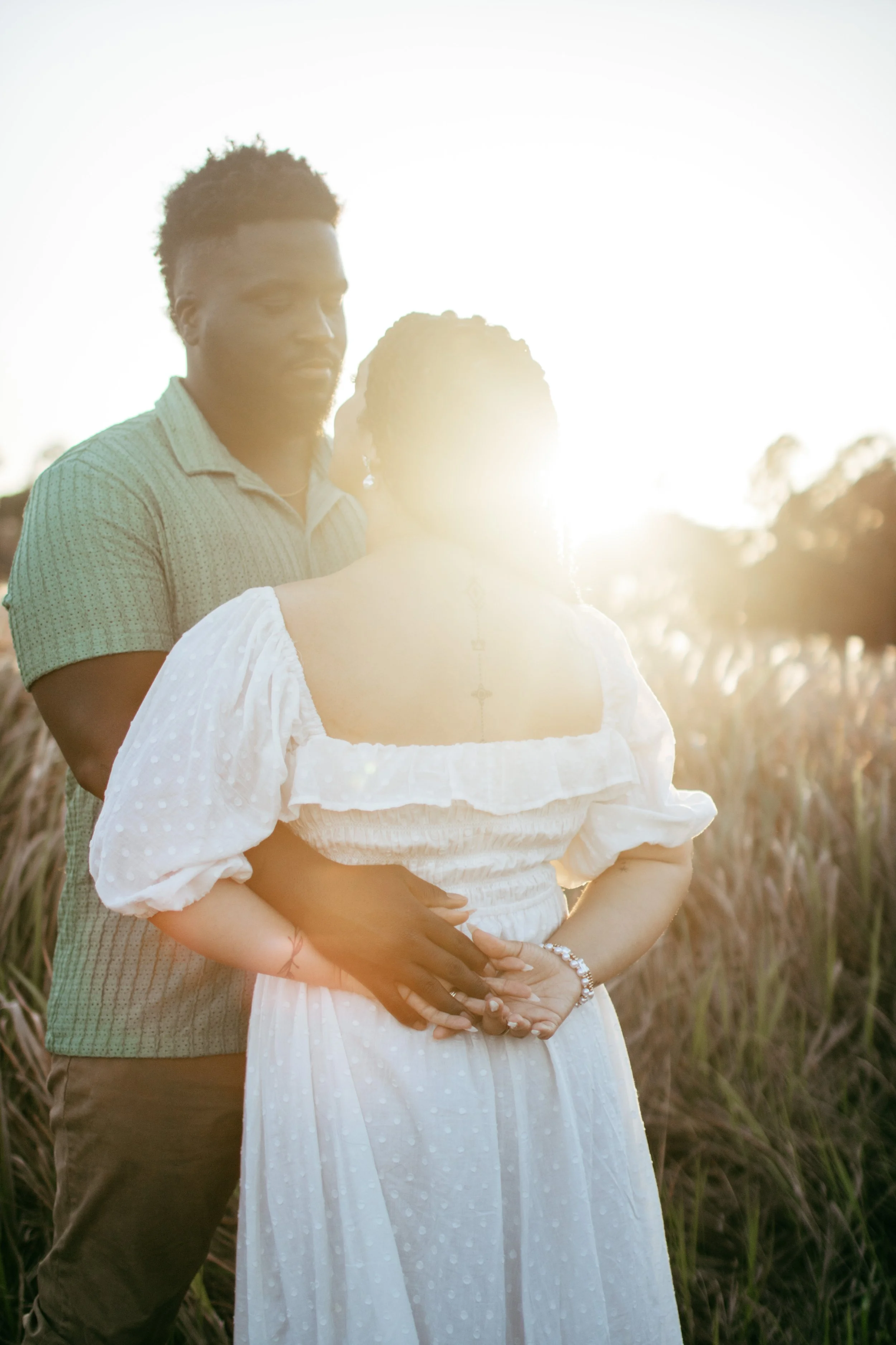 A couple standing in a field at sunset, embracing each other, with the sun behind them creating a warm glow.