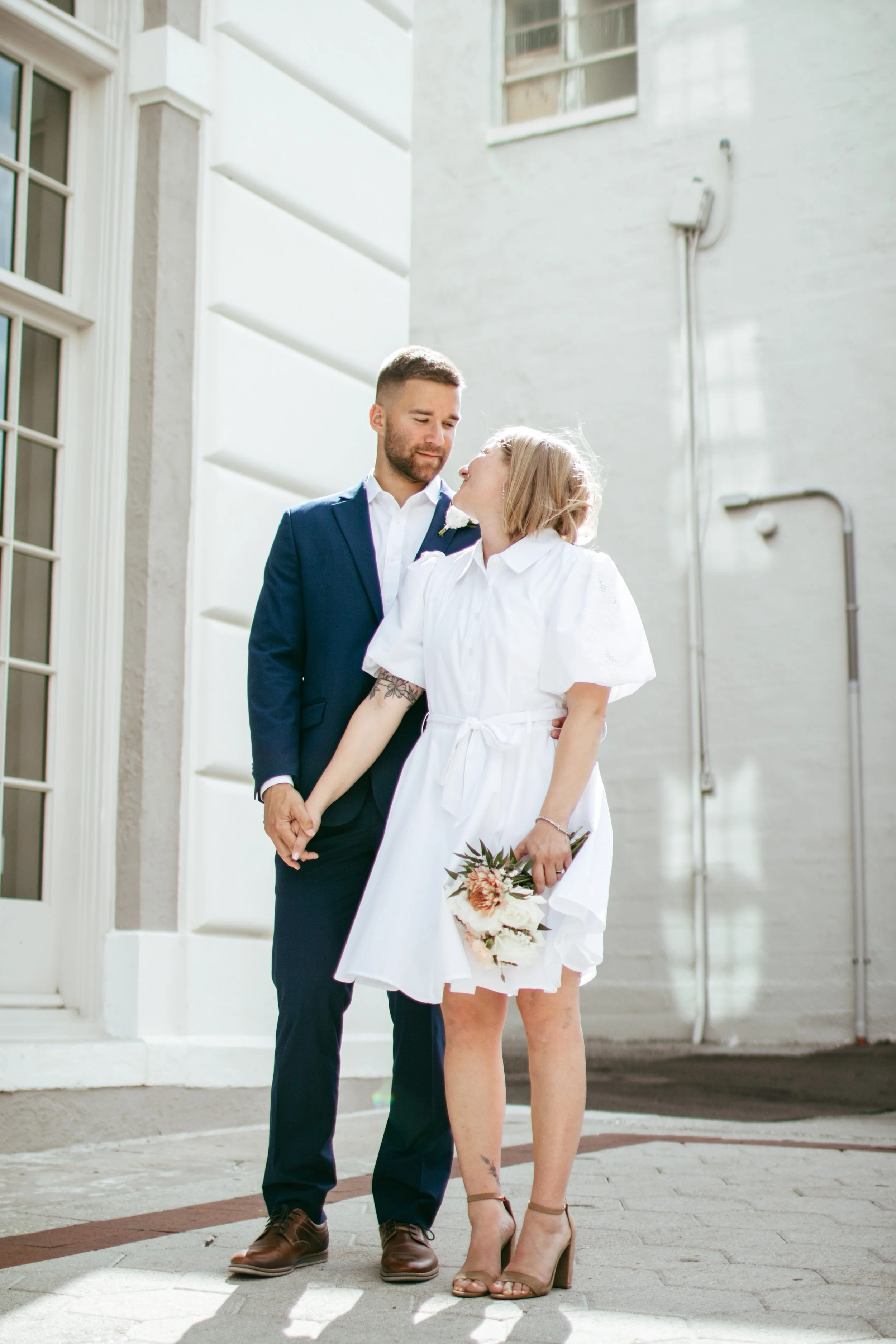 A couple dressed in wedding attire holding hands, with the woman holding a bouquet, standing outside near a white building.