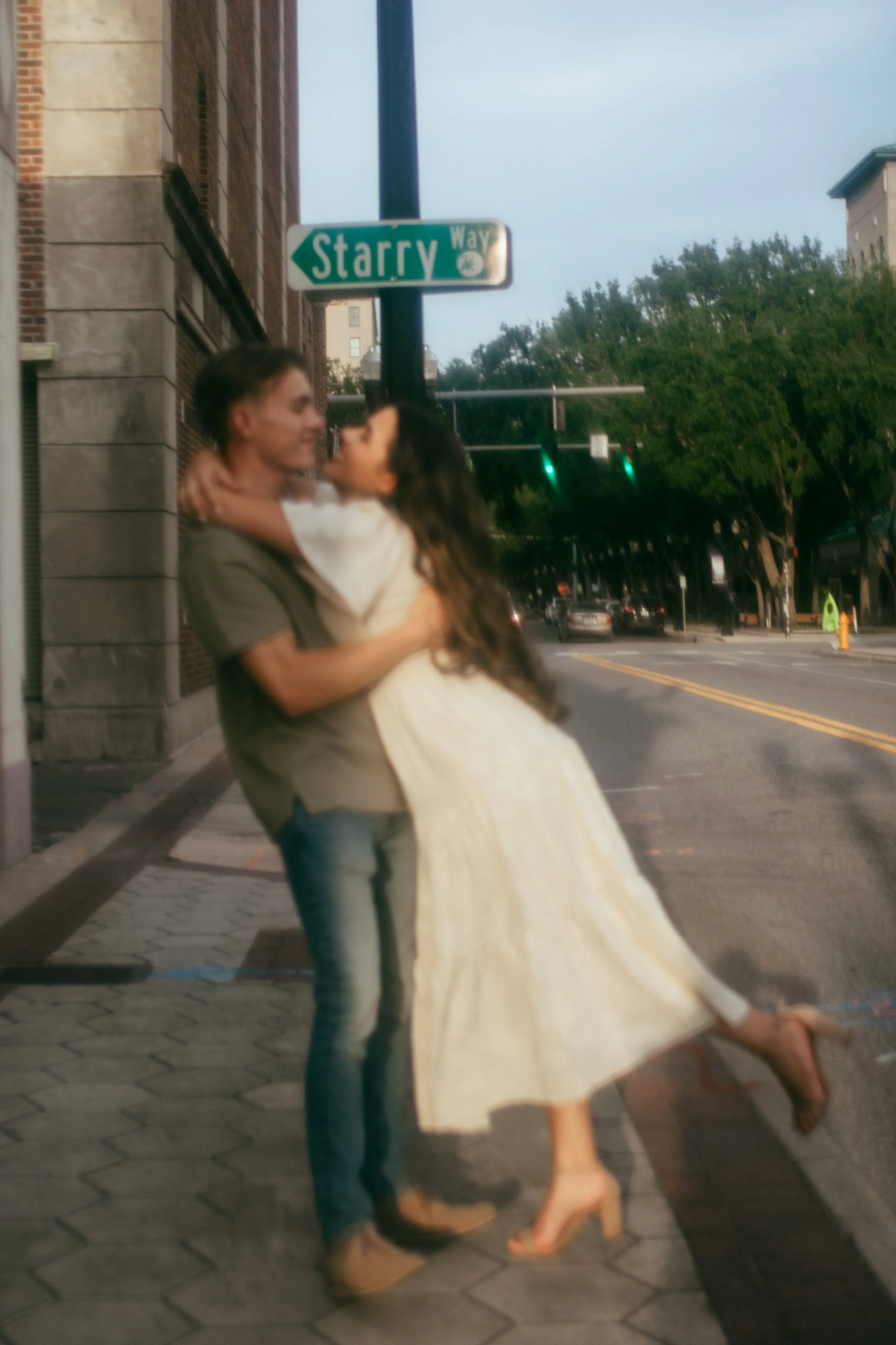 A young couple embracing on a city sidewalk at the corner of Starry Way. The man is holding the woman as she leans back, both smiling affectionately. The woman is wearing a white dress and heels, while the man is dressed casually in a t-shirt and jea