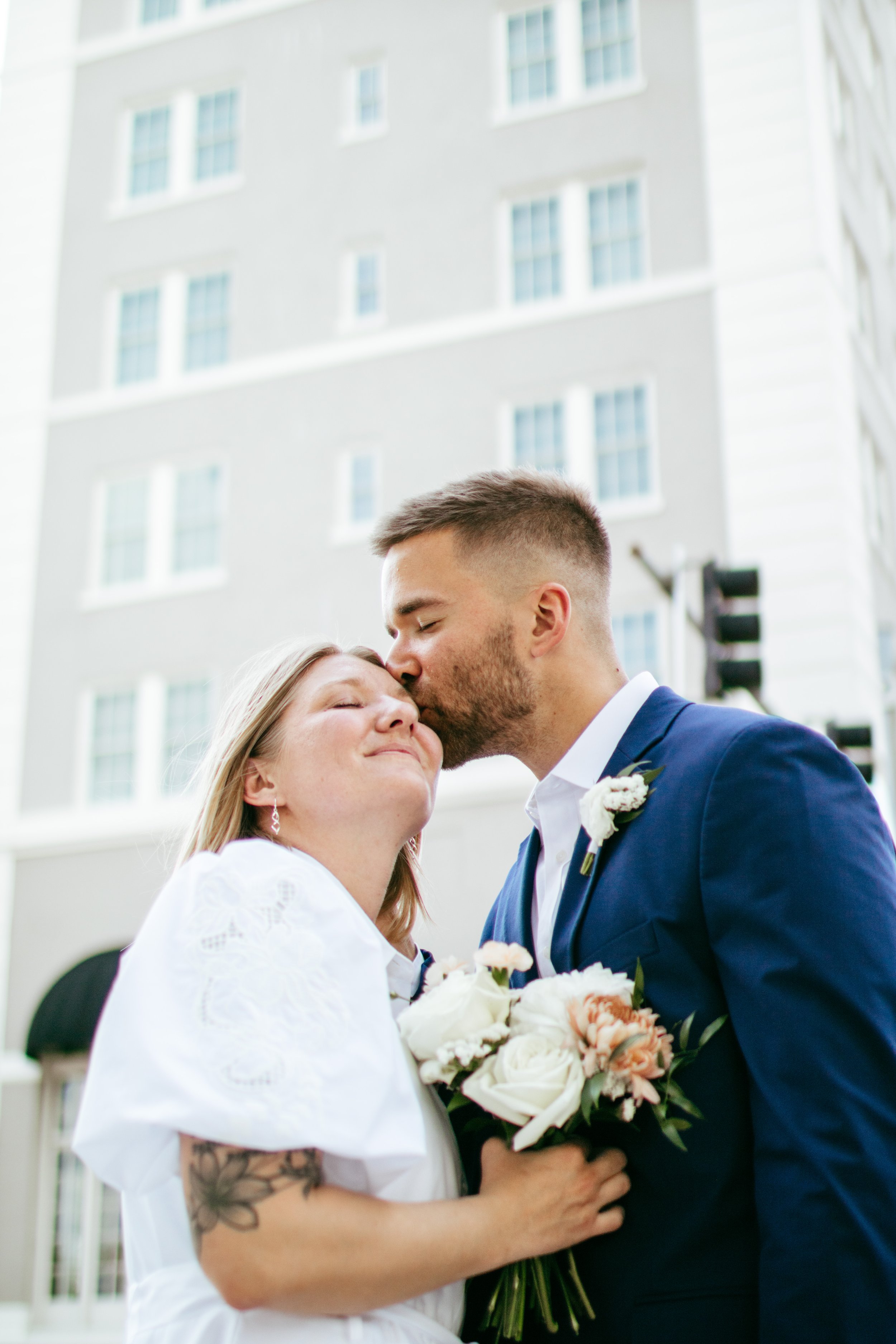 A bride with blonde hair, wearing a white dress and earrings, is smiling with her eyes closed, holding a bouquet of white and blush flowers. A groom with short brown hair and beard, dressed in a navy blue suit and white shirt, is kissing her on the f