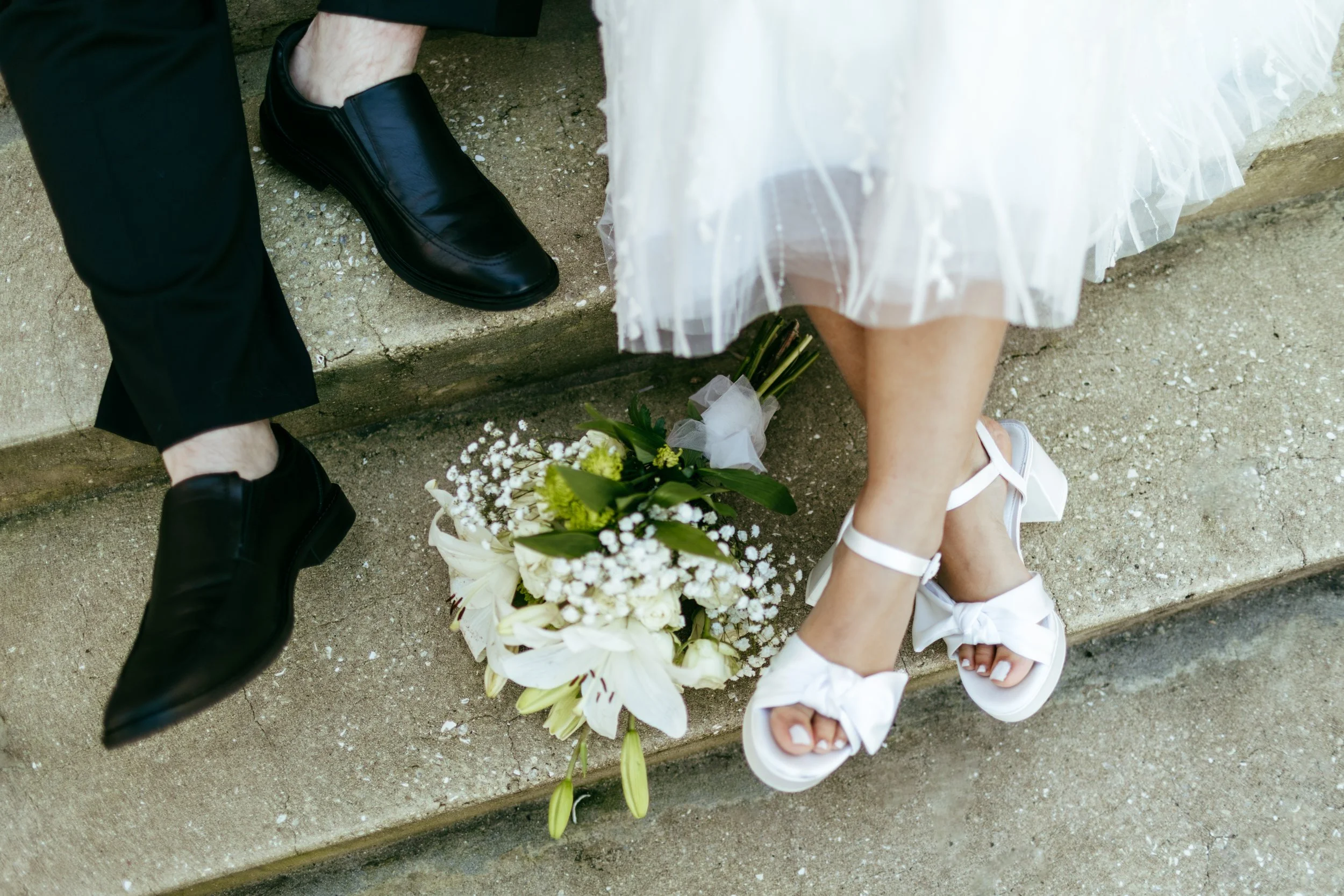 Close-up of two people's legs sitting on concrete stairs, with a bouquet of white flowers between them. One person is wearing black dress shoes and black pants, the other is wearing white sandals and a white tutu skirt.