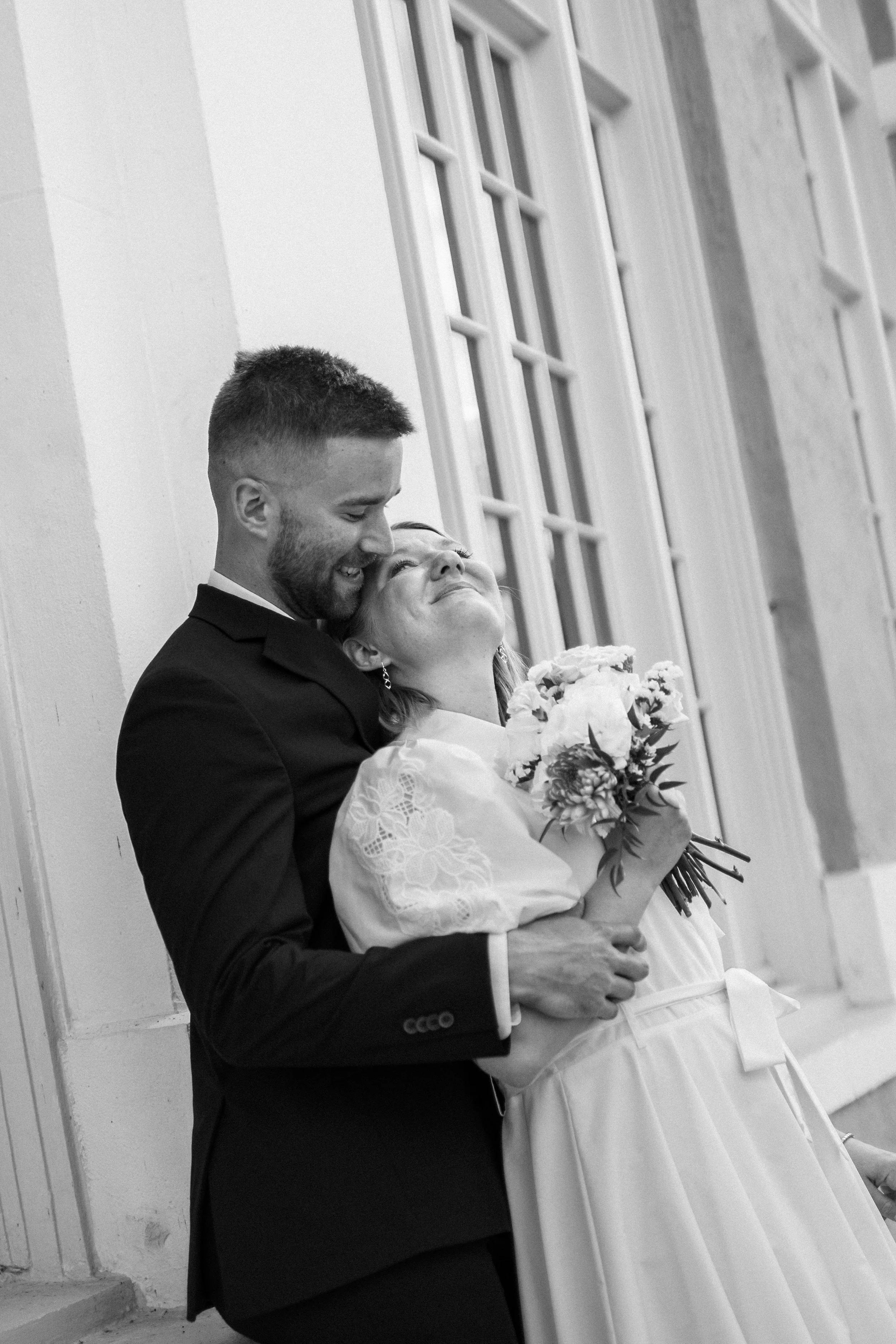 A black and white photo of a smiling couple, a man in a dark suit and a woman in a light dress with a bouquet, sharing a joyful embrace near a large window.