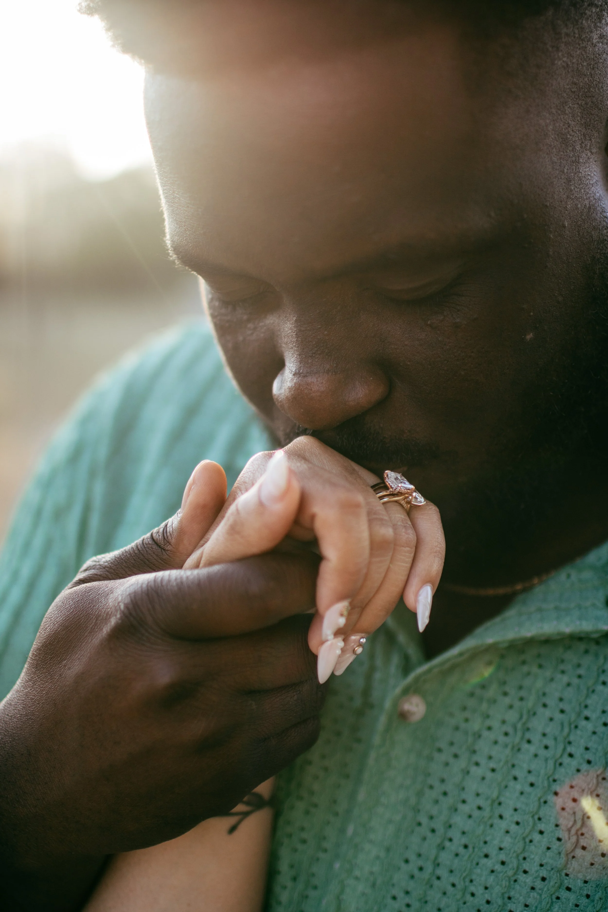 A person with dark skin and long manicured nails is holding their hands together near their mouth, appearing in prayer or deep thought, wearing rings and a green shirt with small patterns, with sunlight softly shining in the background.