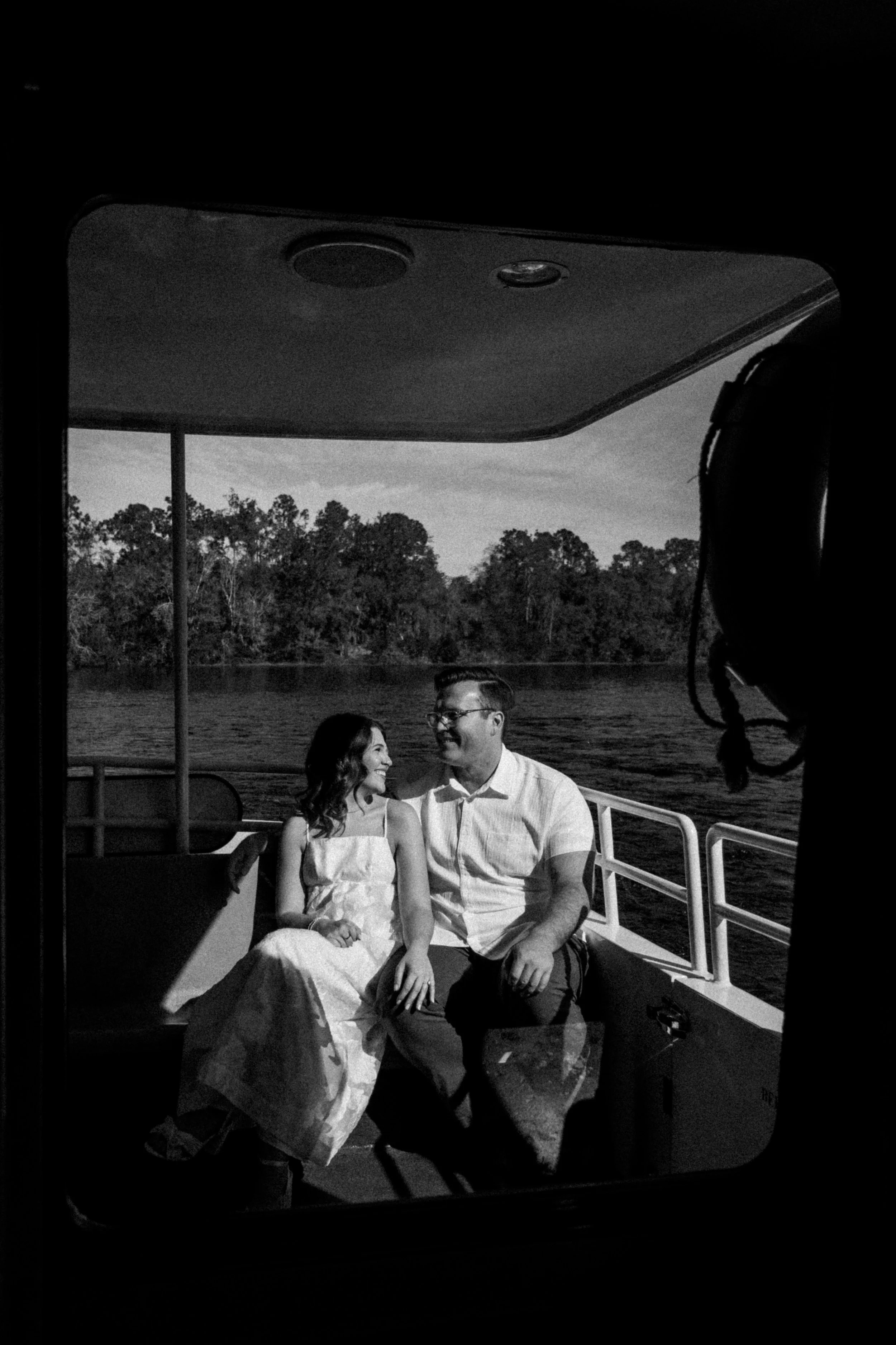 A black and white photo of a couple sitting on a boat, smiling at each other, with trees and water in the background.