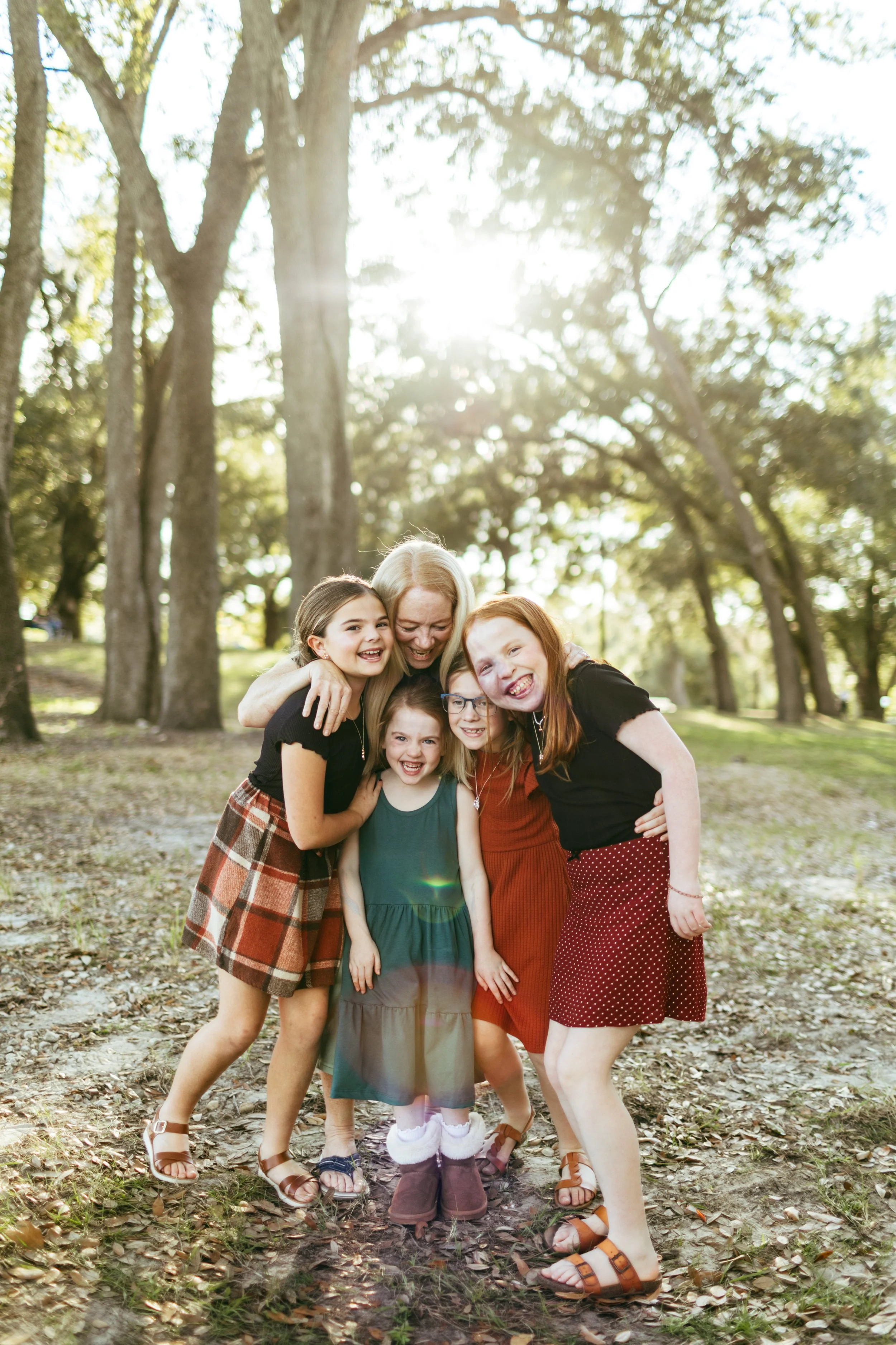 A group of six children, including a woman, hugging and smiling in a park with trees and sunlight in the background.
