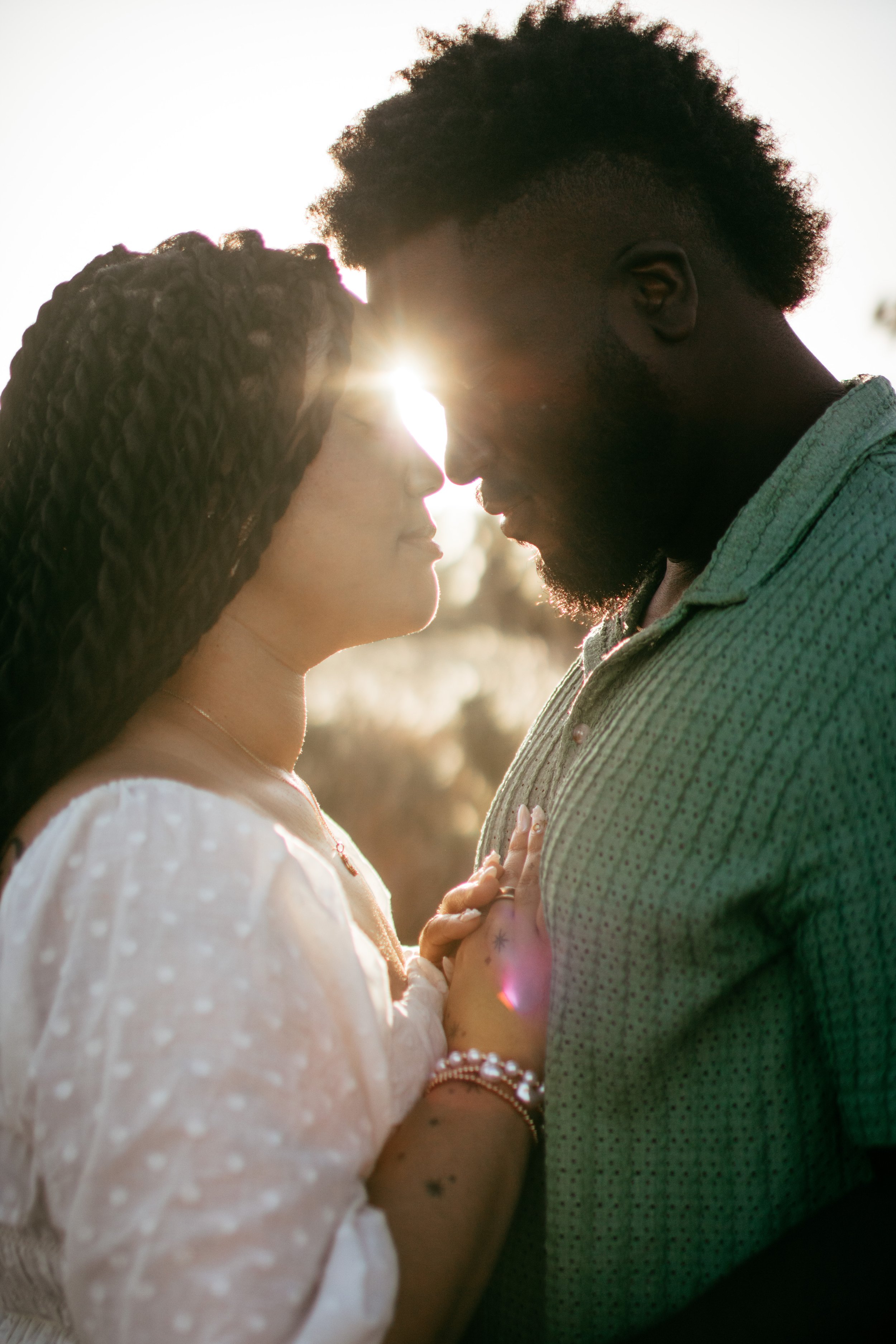 A couple touches foreheads and looks into each other's eyes outdoors during sunset, with sunlight glowing between their faces.
