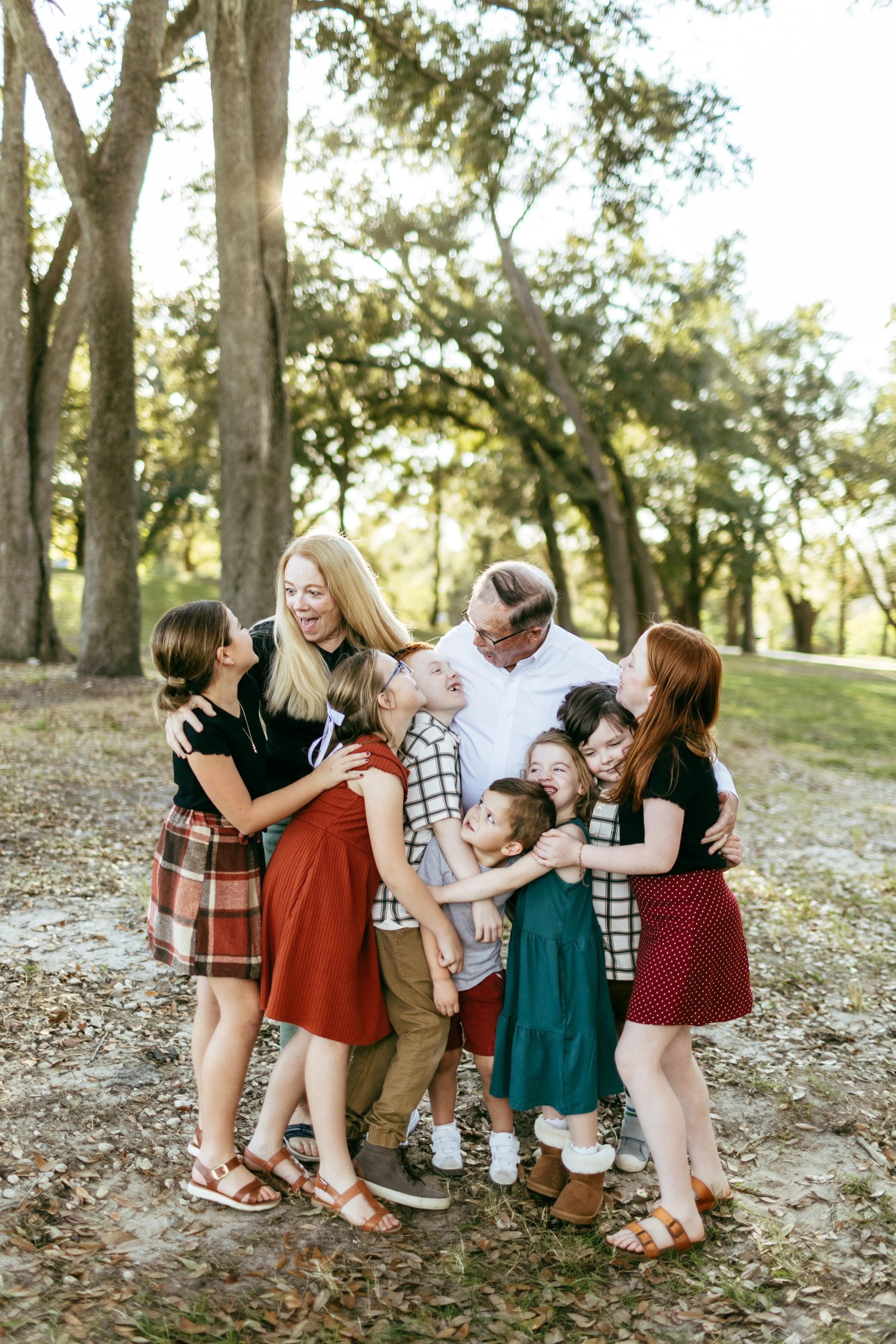 A group of people, including children and adults, embracing and smiling in a park with trees and sunlight in the background.