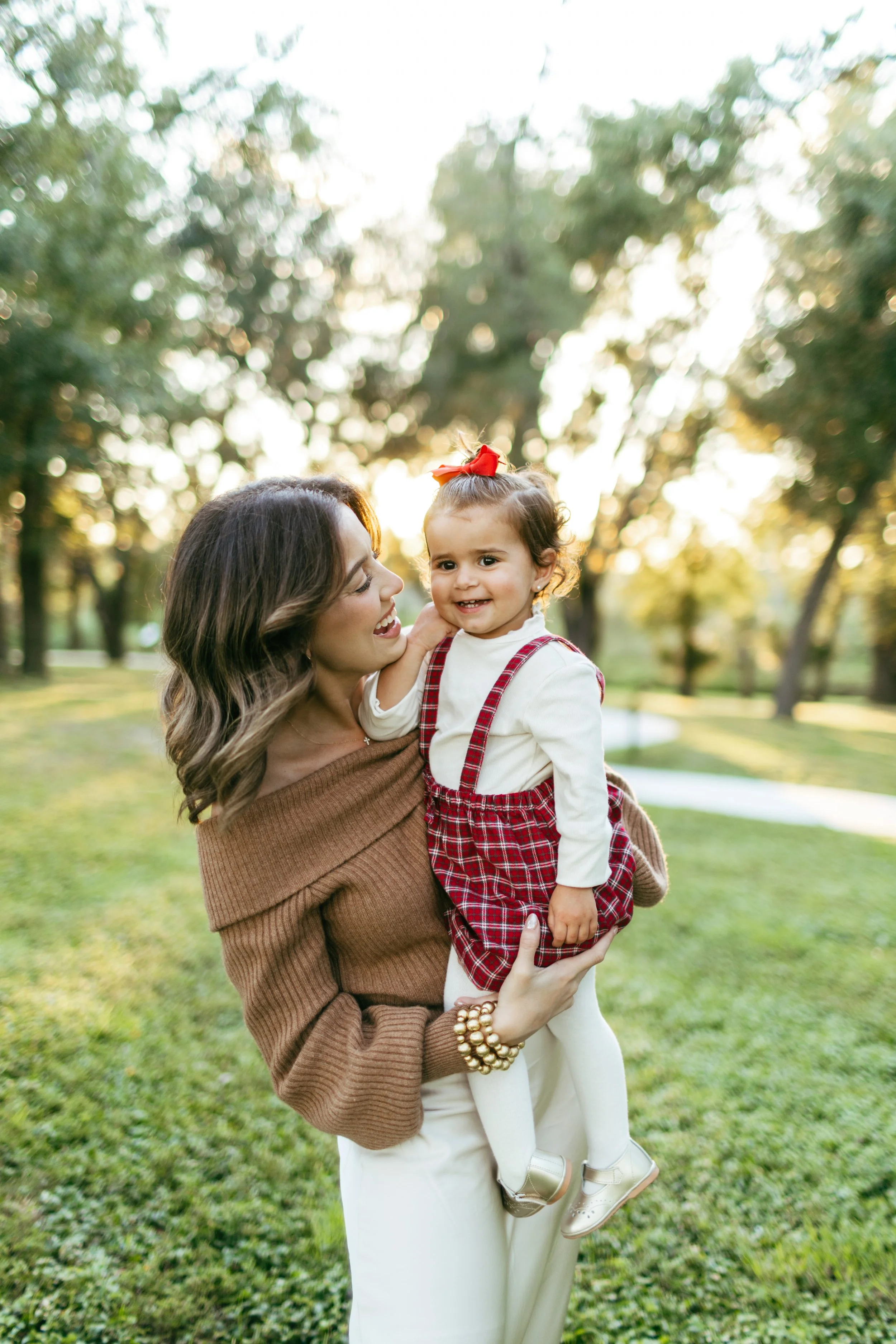 A woman holding a young girl outdoors in a park during sunset, both smiling, with trees and grass in the background.