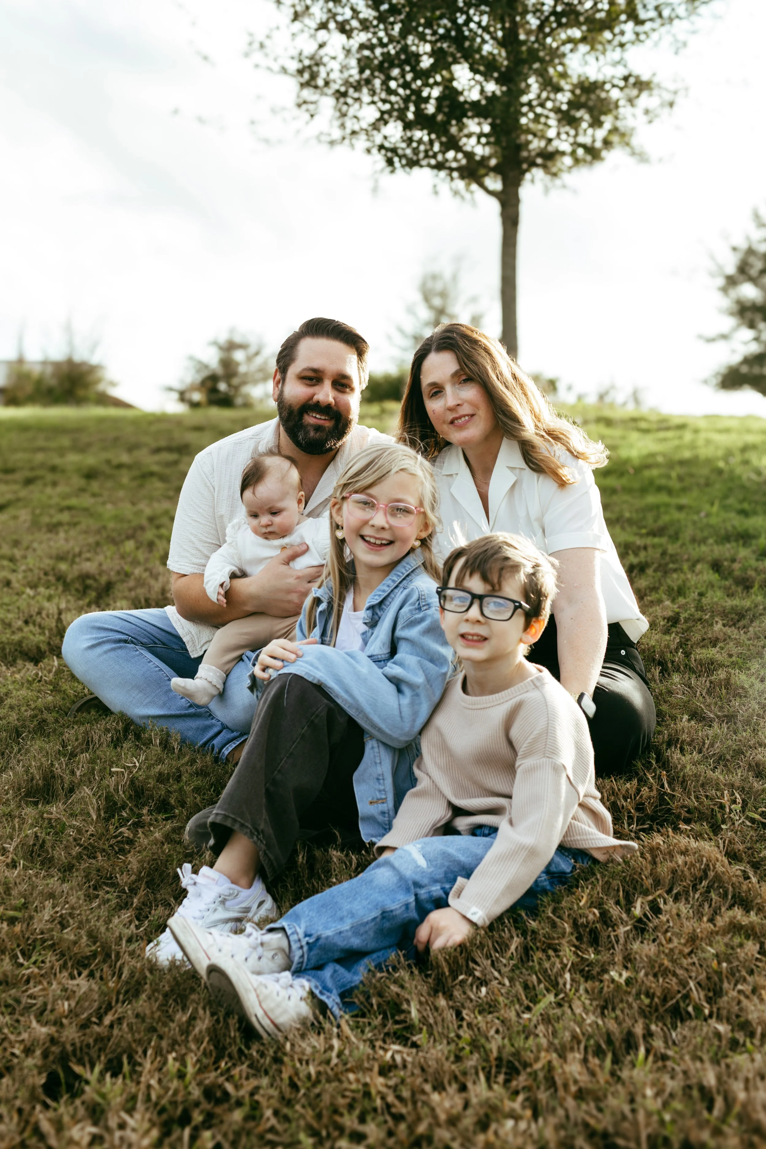 Family of six sitting on grass in park, smiling at camera, with trees and cloudy sky in background