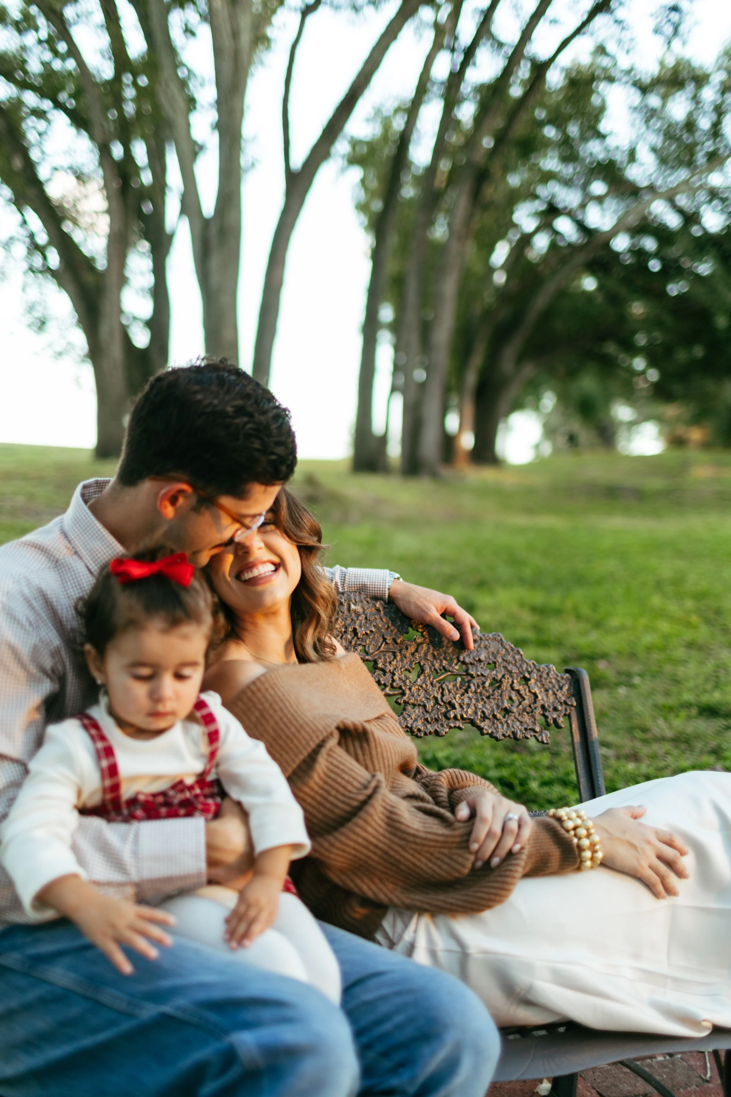 A family of three sitting on a park bench, smiling and enjoying each other's company during the daytime, with trees in the background.