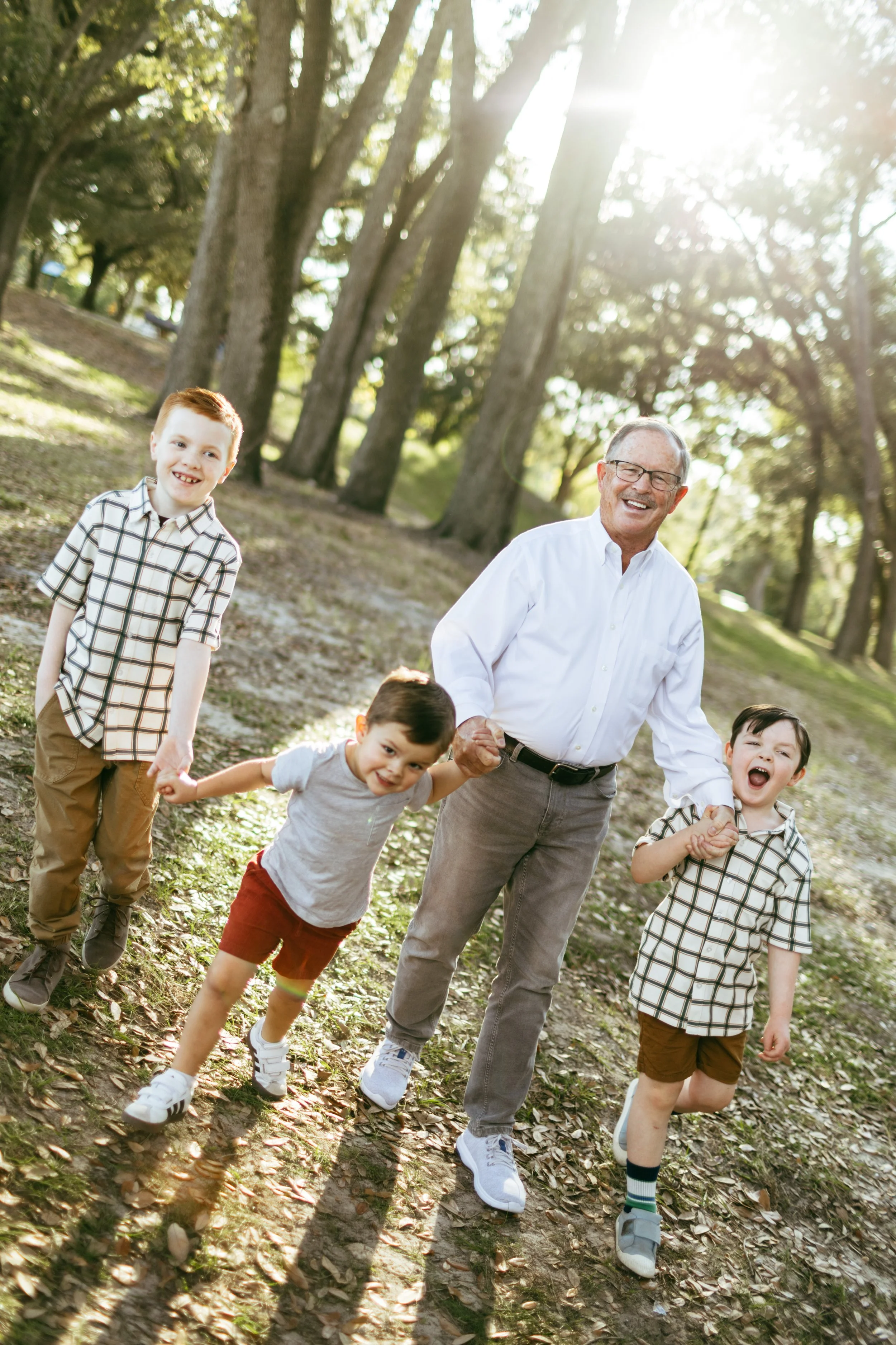 An elderly man smiling and holding hands with three young boys while walking through a wooded park with sunlight filtering through the trees.