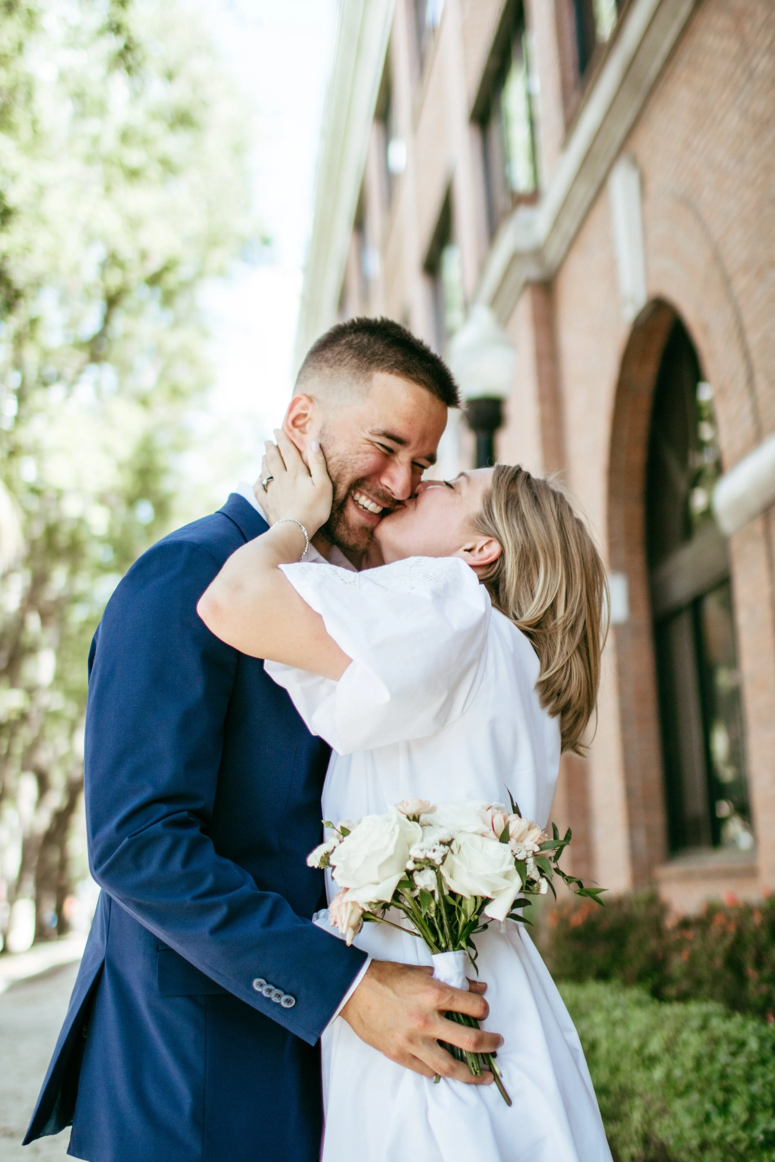 A couple sharing a kiss outdoors, the woman holding a bouquet of white flowers, with trees and a brick building in the background.