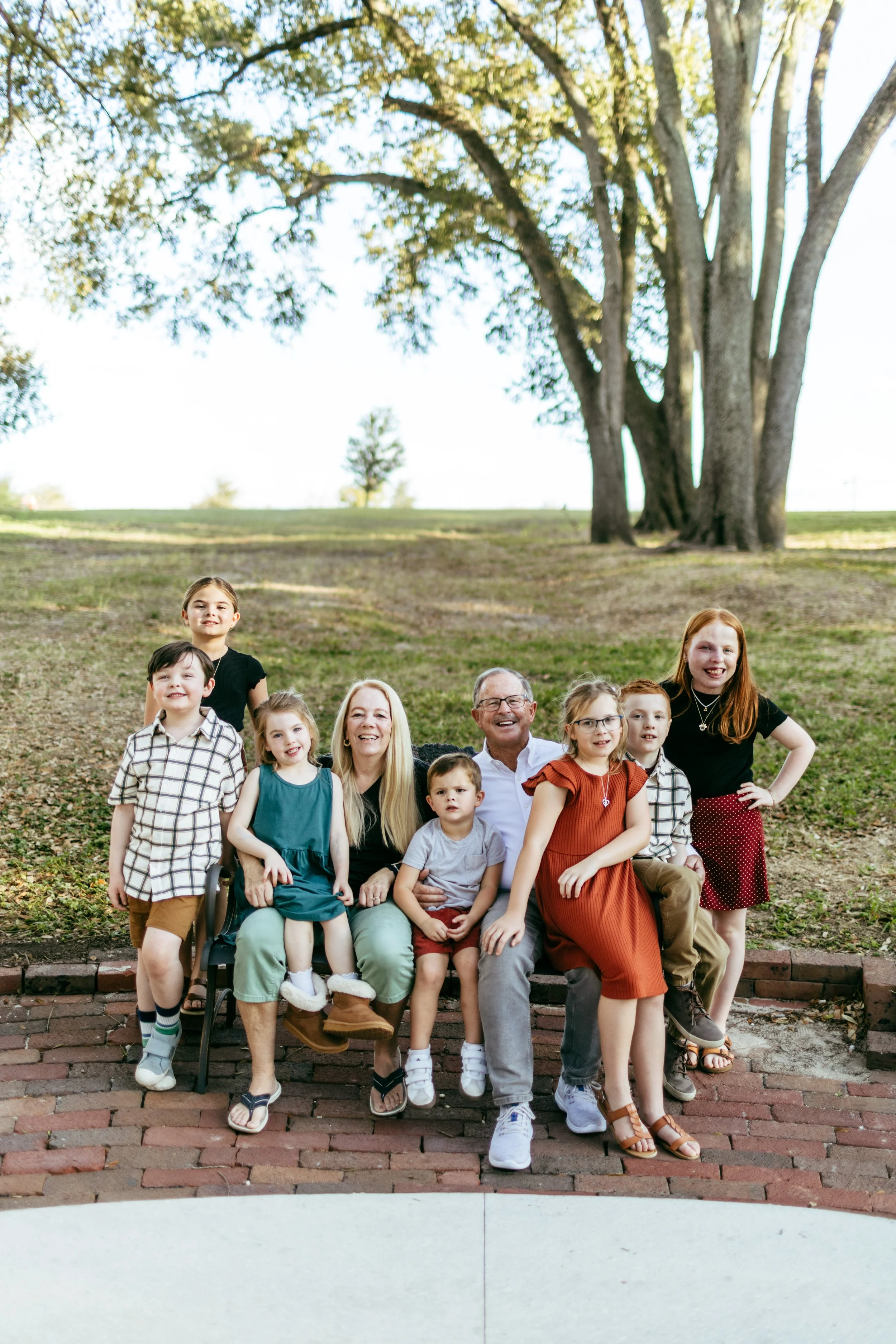A multigenerational family group portrait outdoors on a brick pathway with grass and large trees in the background, featuring adults and children smiling and sitting together.