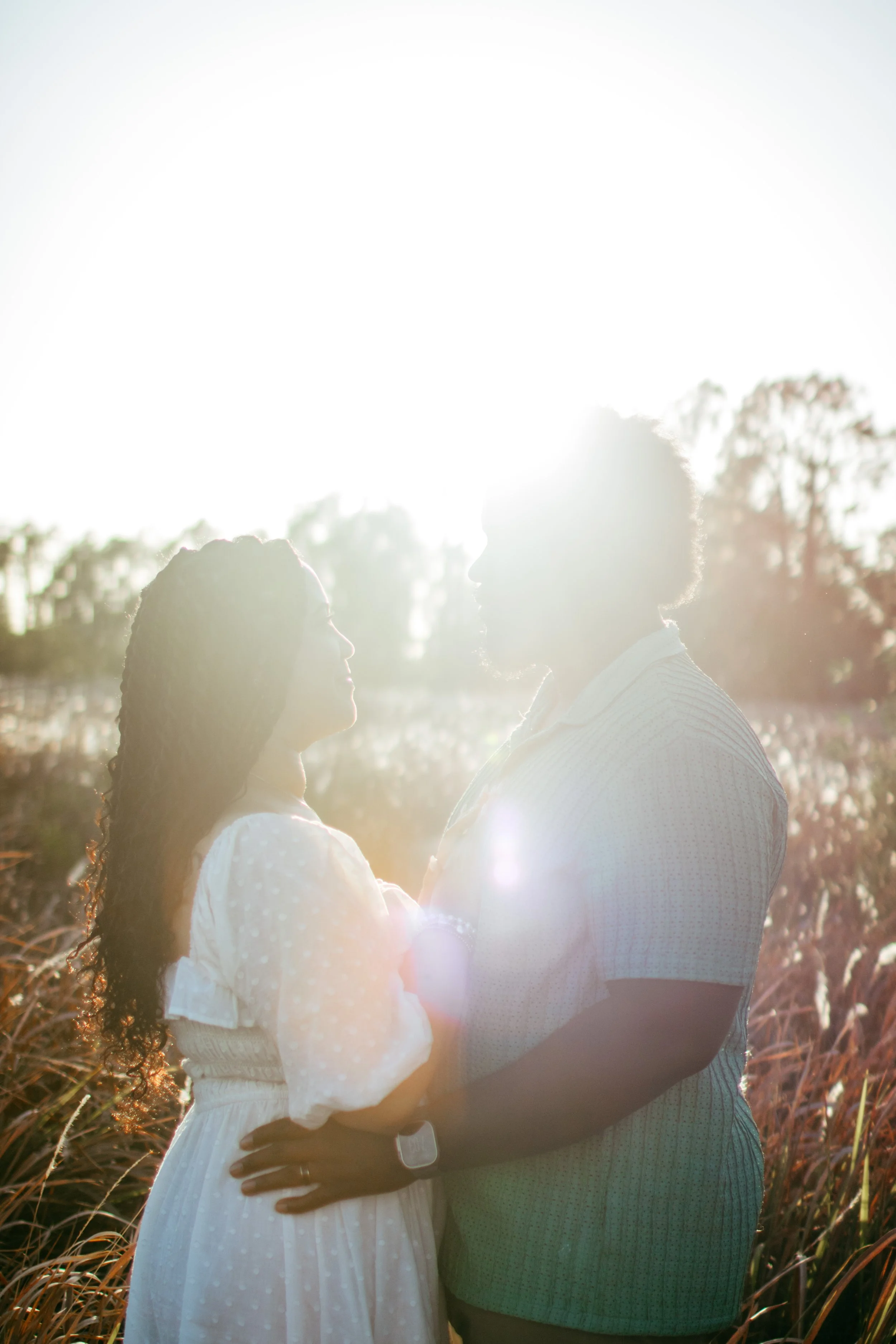 A couple standing outdoors in a field at sunset, facing each other and holding hands, with sunlight making them appear as silhouettes.