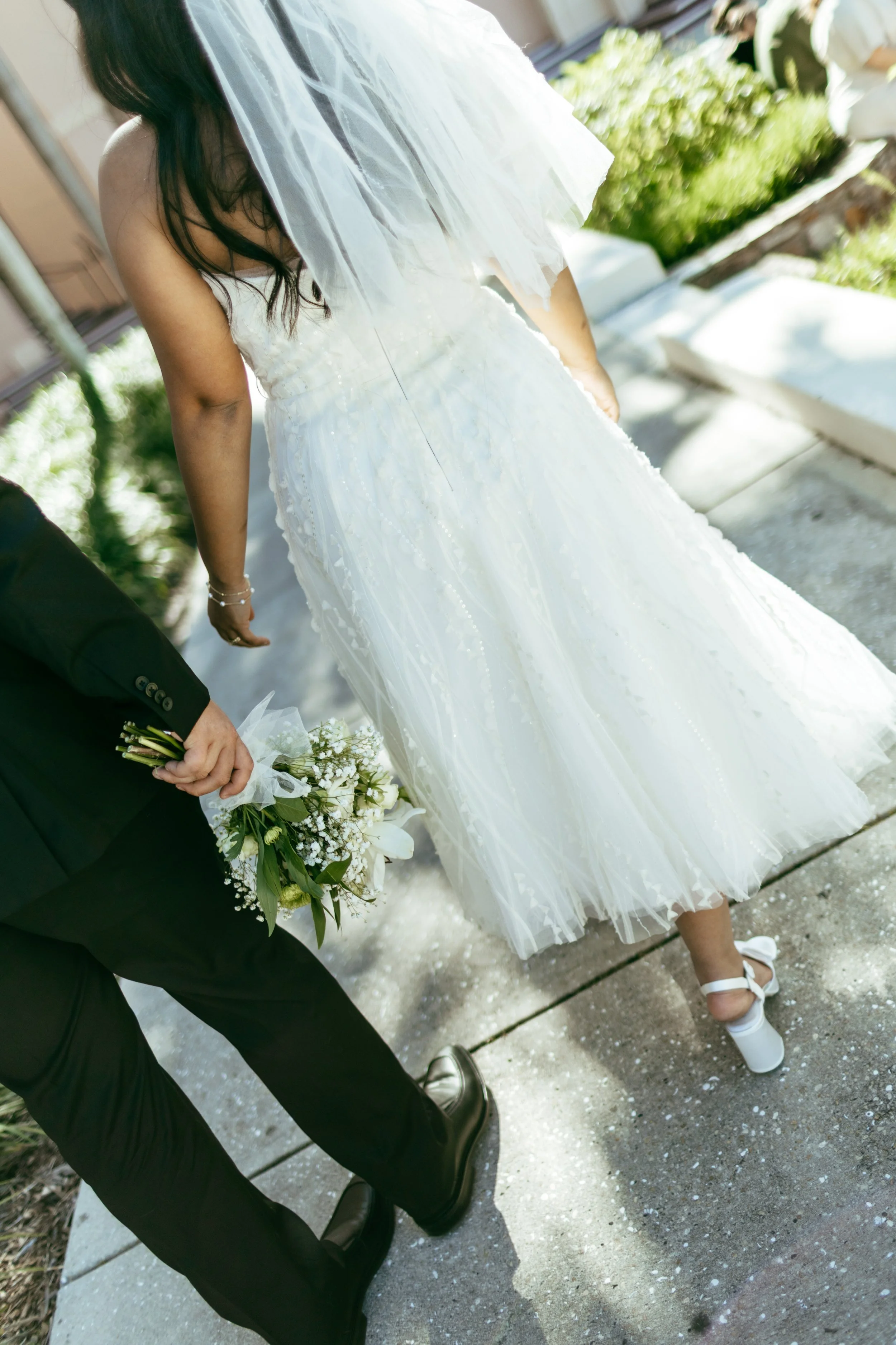 Bride in a white wedding dress walking outdoors, holding a bouquet, with a person in a black suit.