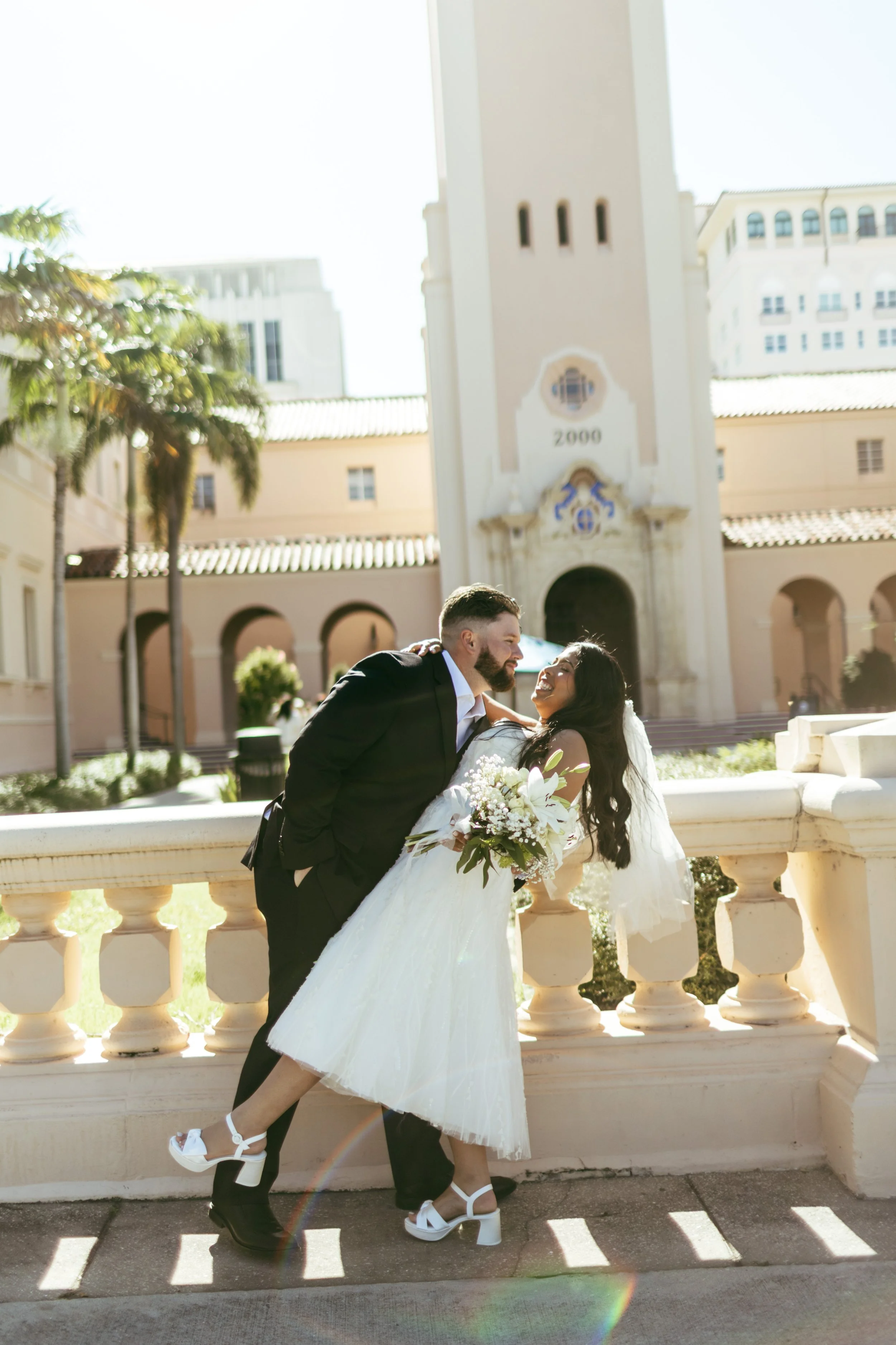 A bride and groom share a kiss outdoors near a historic building, with palm trees and bright sunlight in the background.