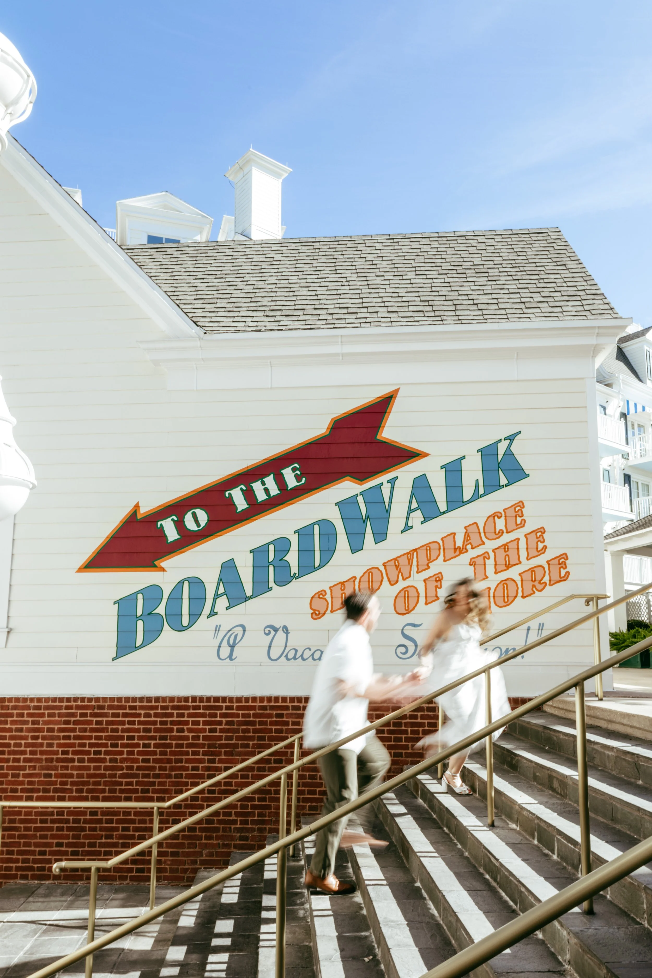People walking up outdoor stairs past a large painted sign on a white building that reads 'To the Boardwalk Showplace of the Shore' with a red arrow pointing to the right, under a clear blue sky.