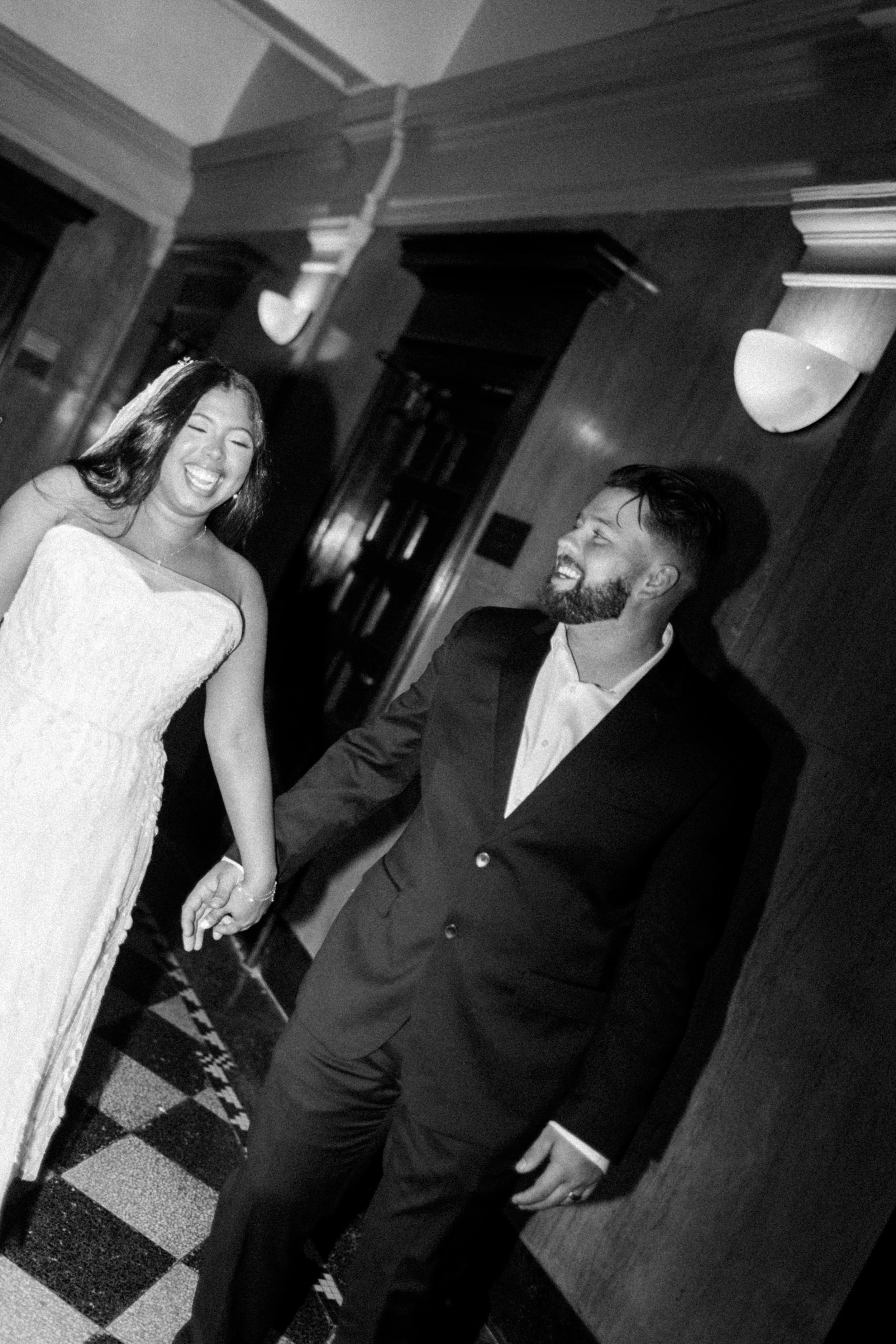 A woman in a strapless wedding dress and a man in a suit holding hands and smiling at each other in an indoor setting.