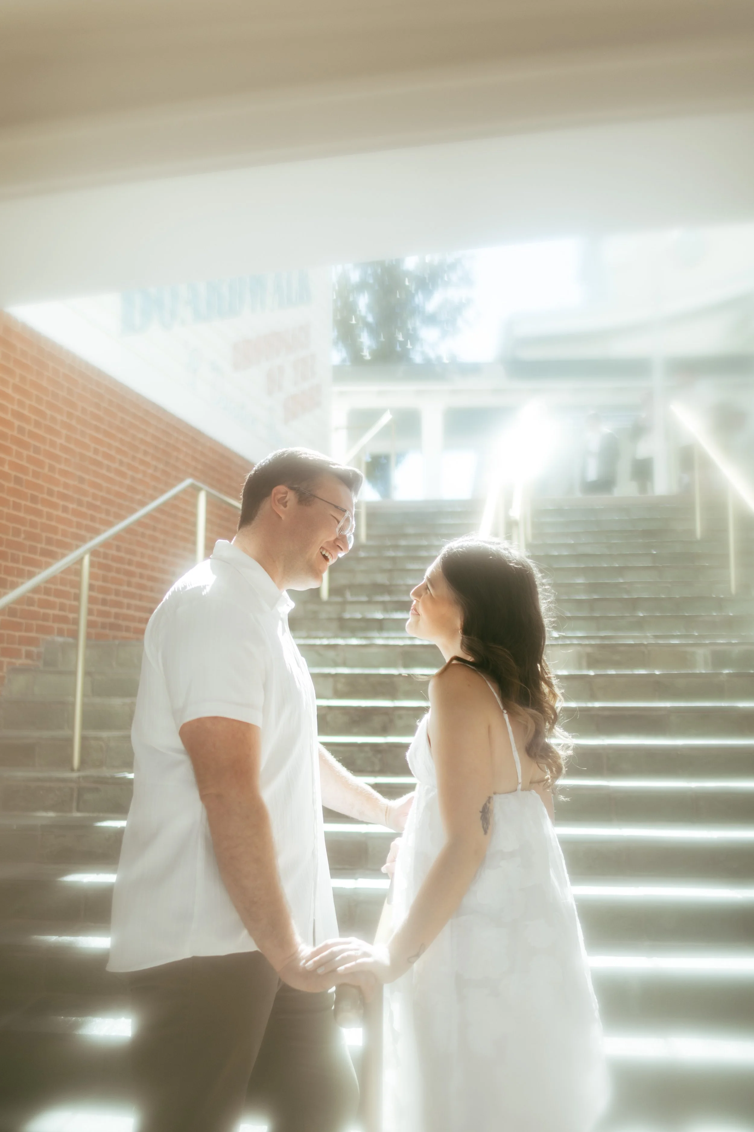 A couple holding hands and smiling at each other in front of a sunlit staircase, with a brick wall on one side and bright sunlight shining from the top of the stairs.