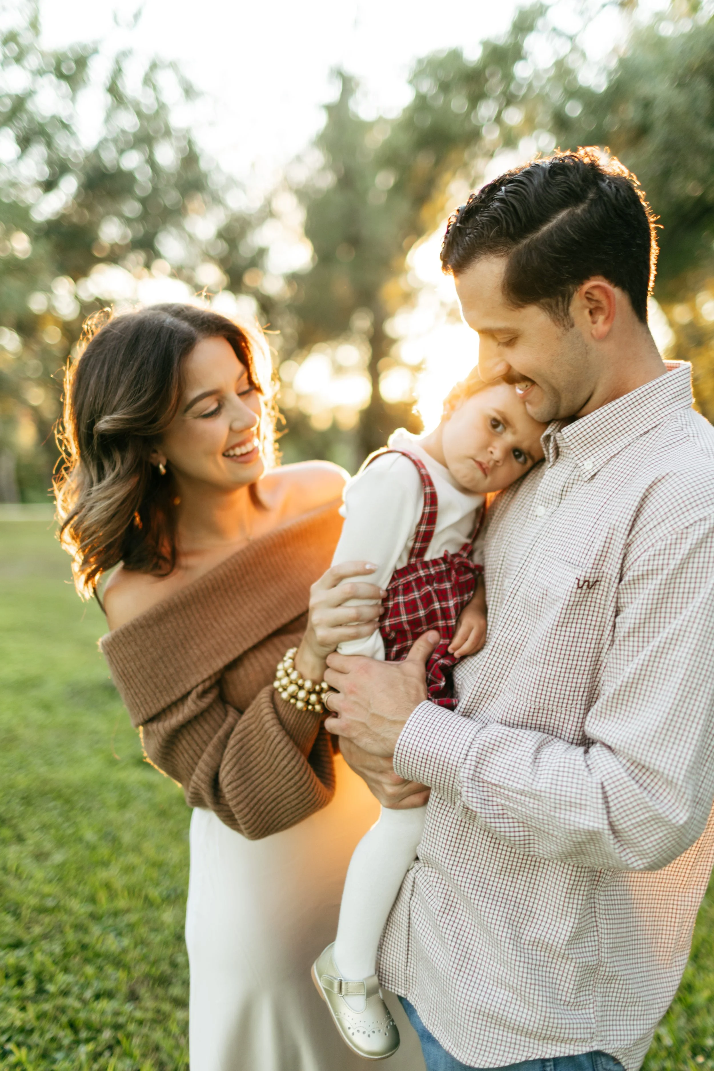 A family of three stands outdoors in a park at sunset; the mother is smiling at the father, who is holding their young daughter close, with sunlight glowing around them.