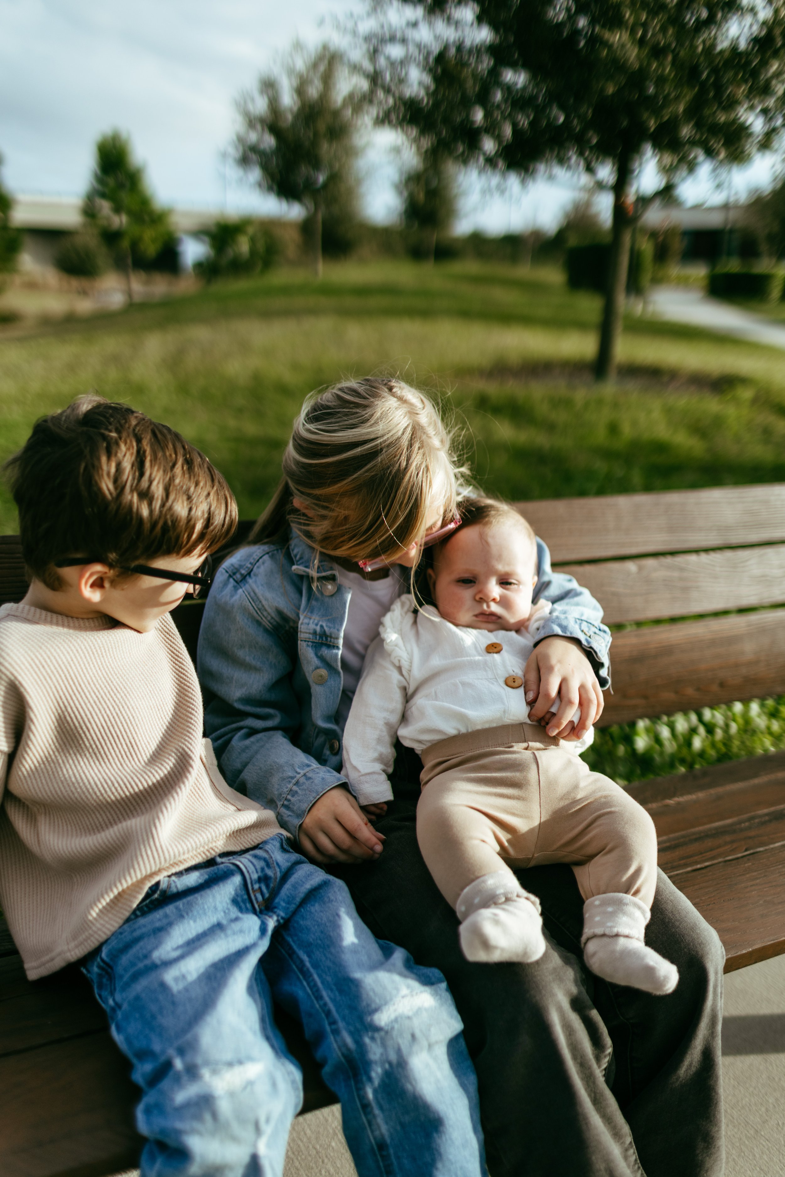 A woman with two young children sitting on a park bench outdoors. The woman is holding a baby and a young boy sitting beside her.
