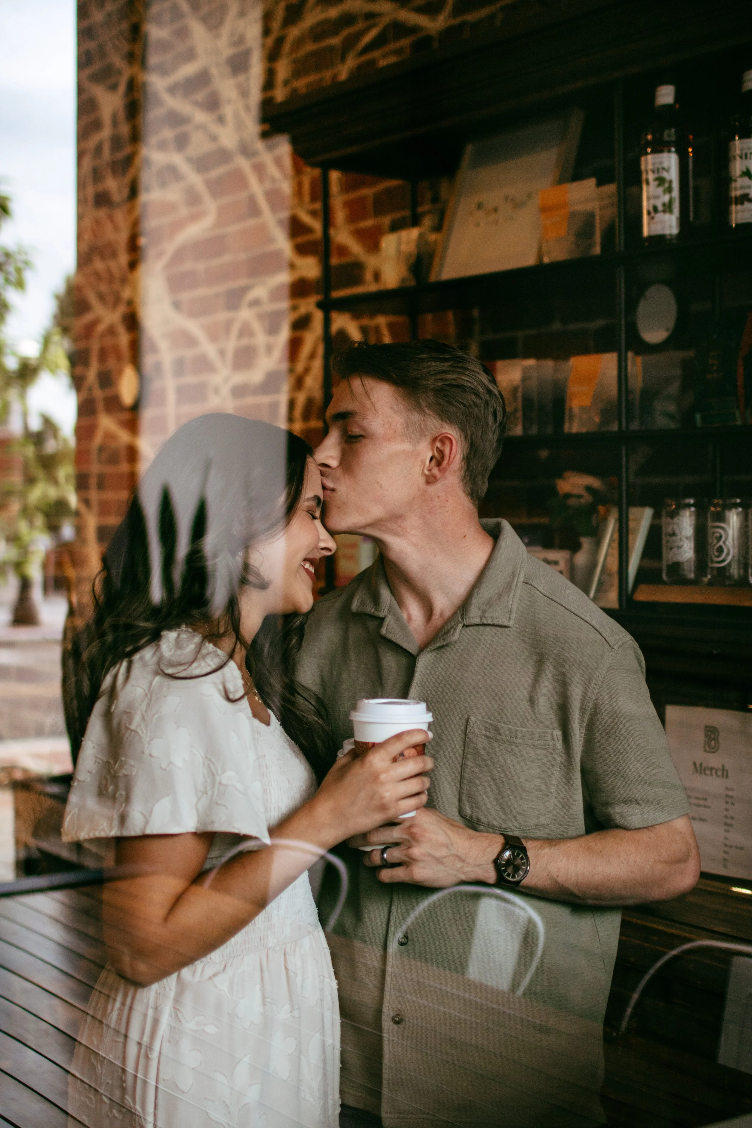 A couple sharing an intimate moment inside a cafe, with the man kissing the woman's forehead while she holds a coffee cup, seen through a glass window.