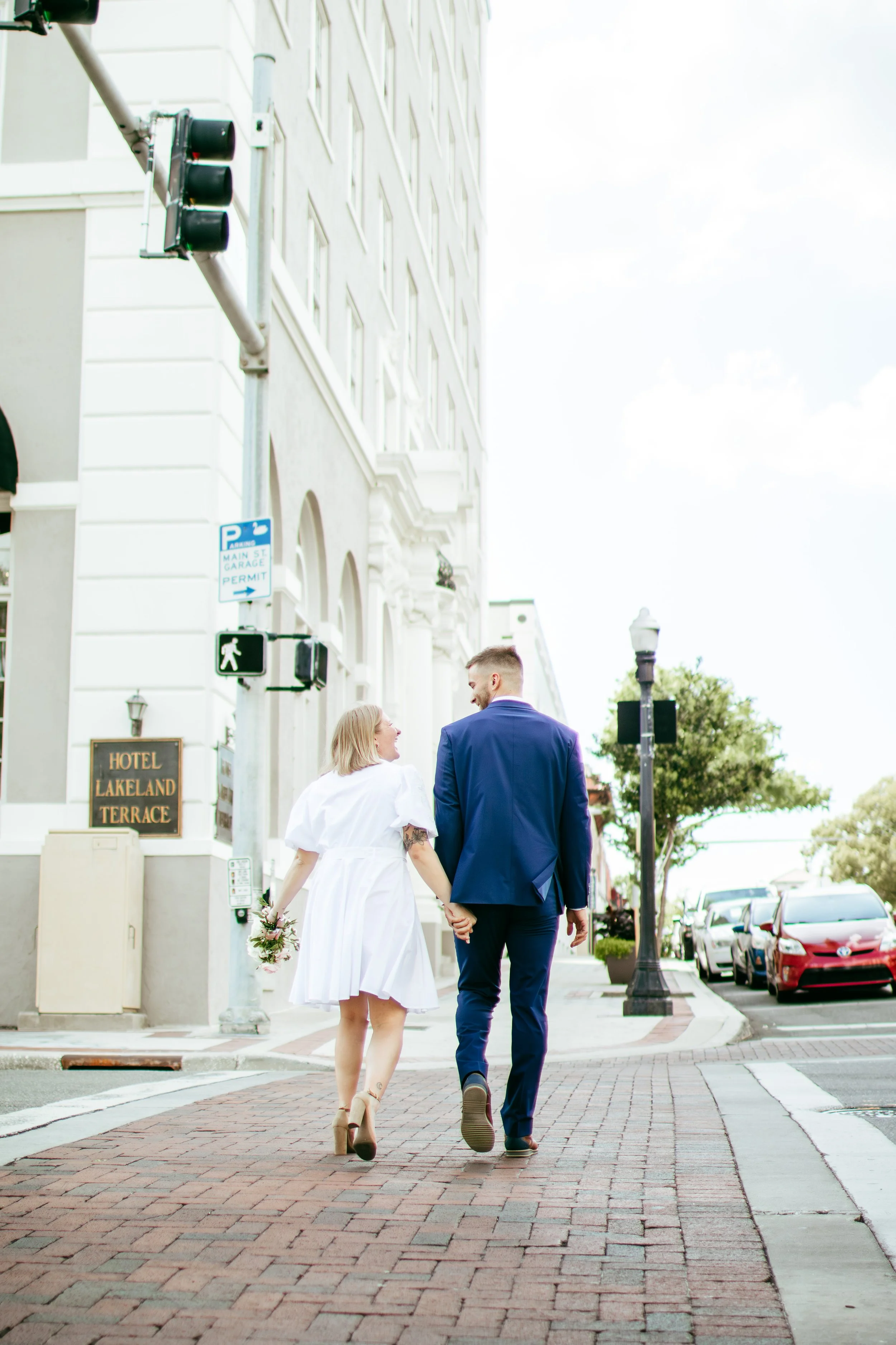 A couple walking hand in hand in an urban setting, with the woman holding a small bouquet, on a brick sidewalk near a white building with a hotel sign.