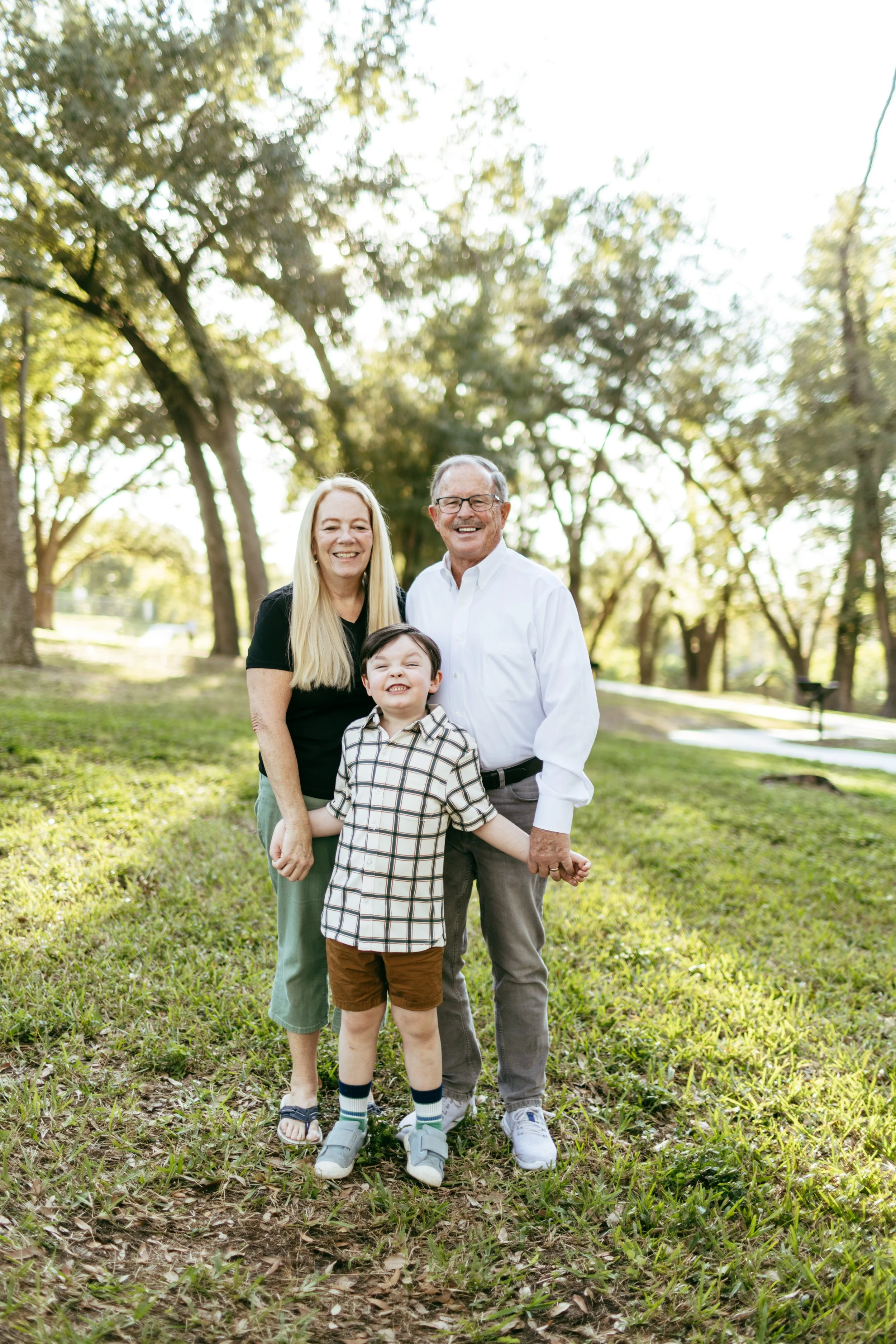 A family of three with a man, woman, and young boy holding hands outdoors in a park, smiling at the camera on a sunny day.