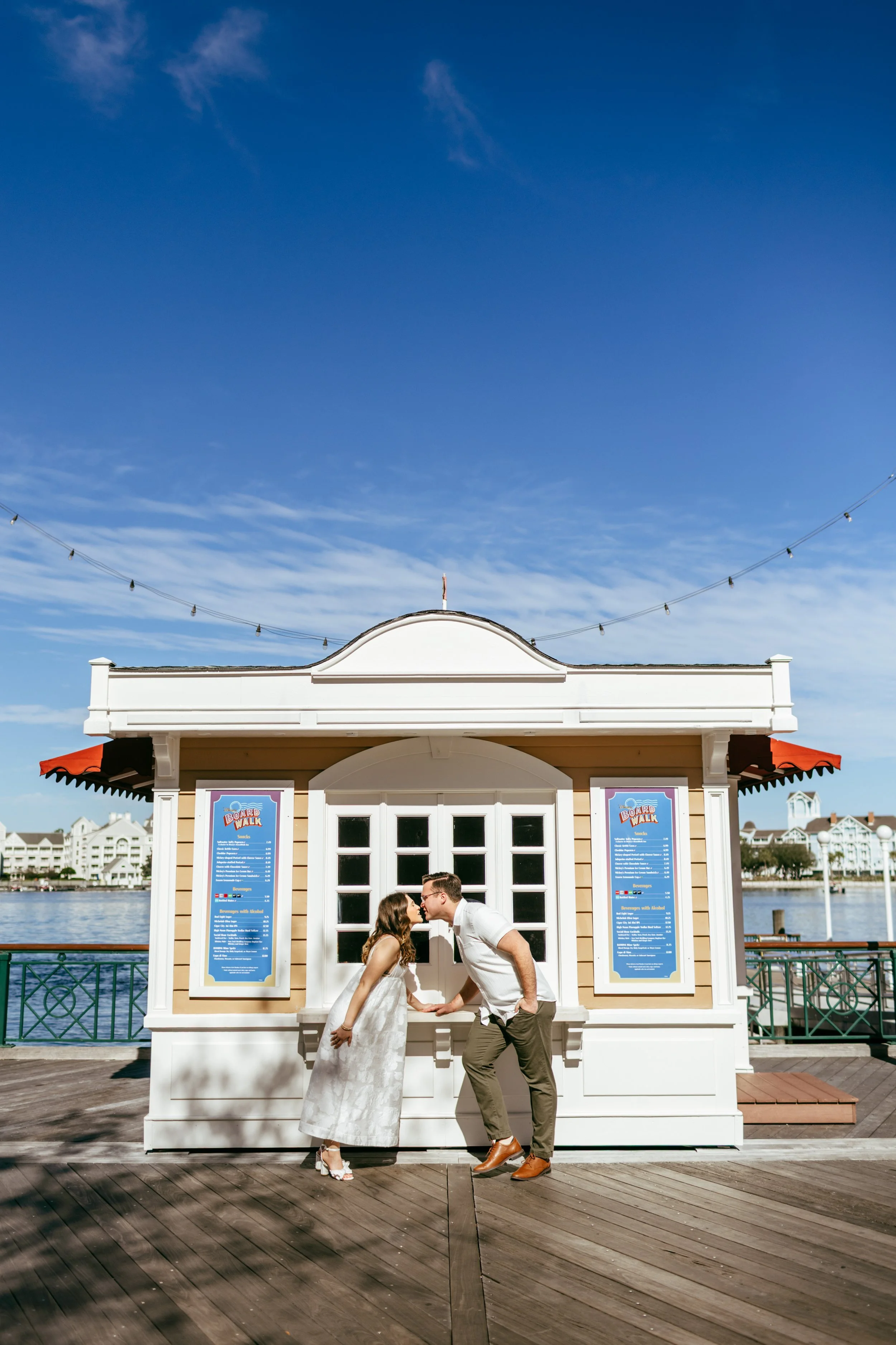 A couple kissing in front of a small building on a wooden dock near water, with buildings and blue sky in the background.