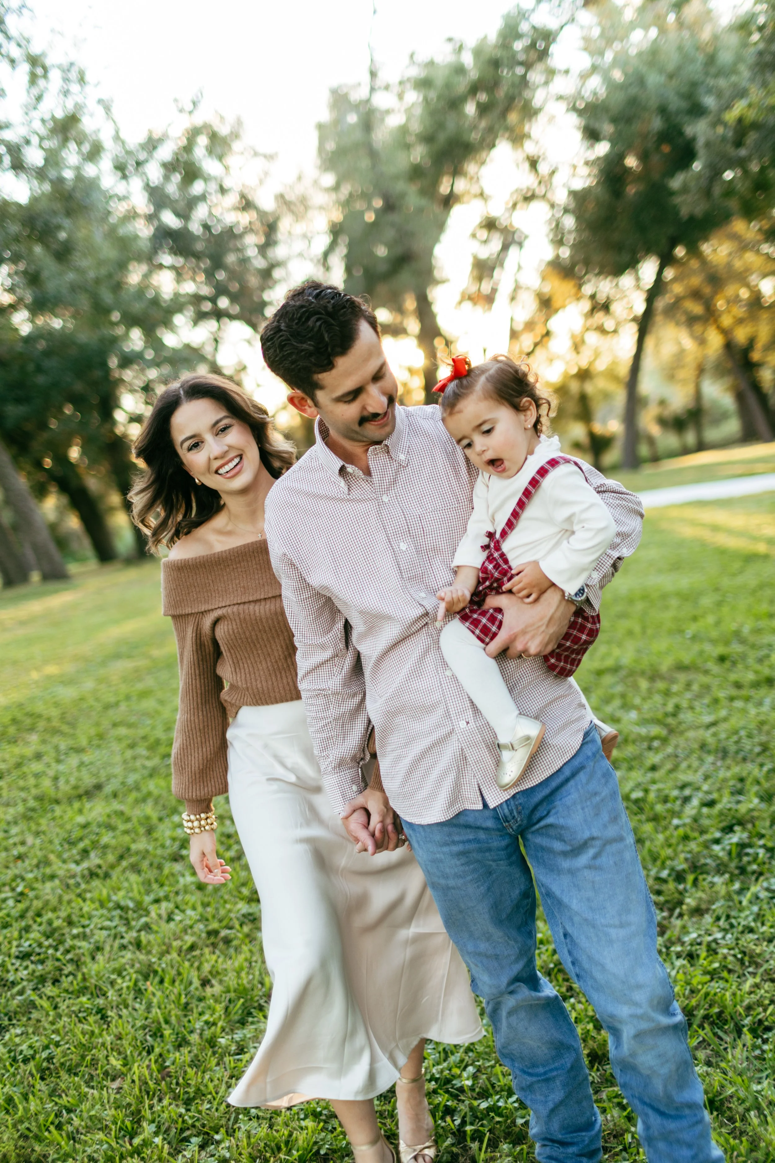 A happy family of three walking outdoors in a park during sunset. The father is holding a young girl, and the mother is walking beside them, all smiling and enjoying the moment.