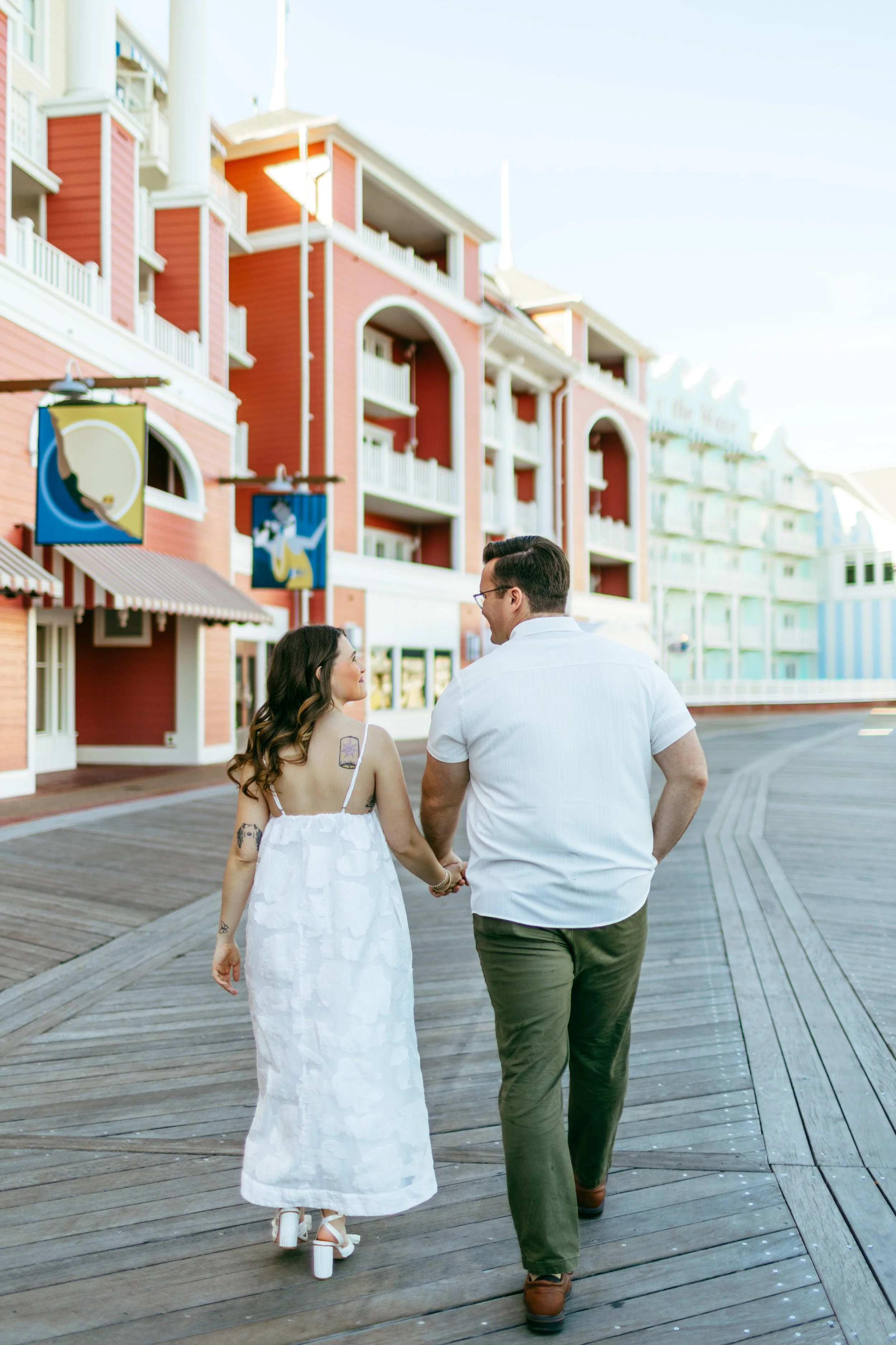 A couple holding hands and walking on a wooden boardwalk in a seaside town, with colorful buildings and beach-themed signs in the background.