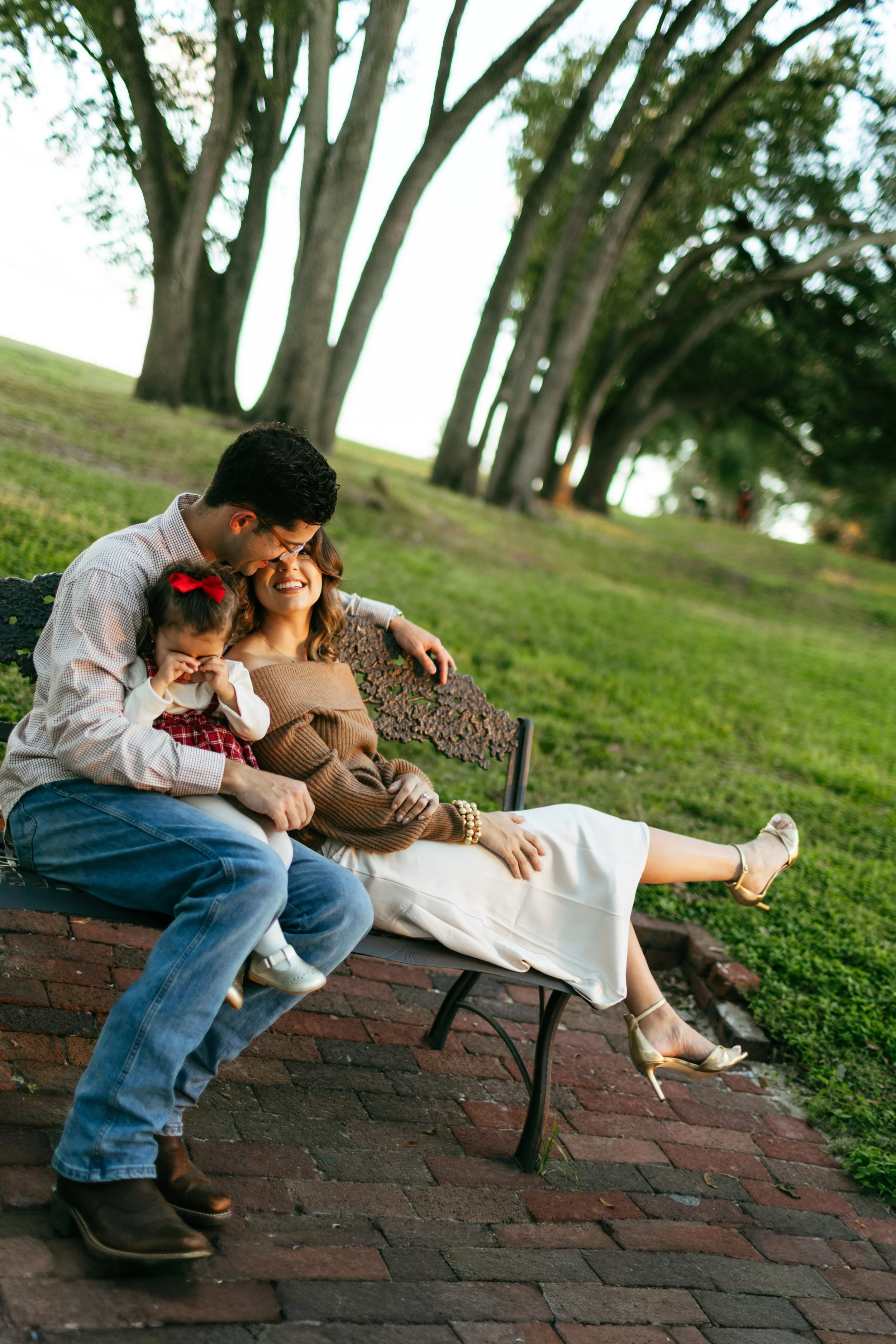 A family of three sitting on a park bench outdoors during the daytime, with tall trees in the background. The father, mother, and young daughter are laughing and enjoying each other's company.