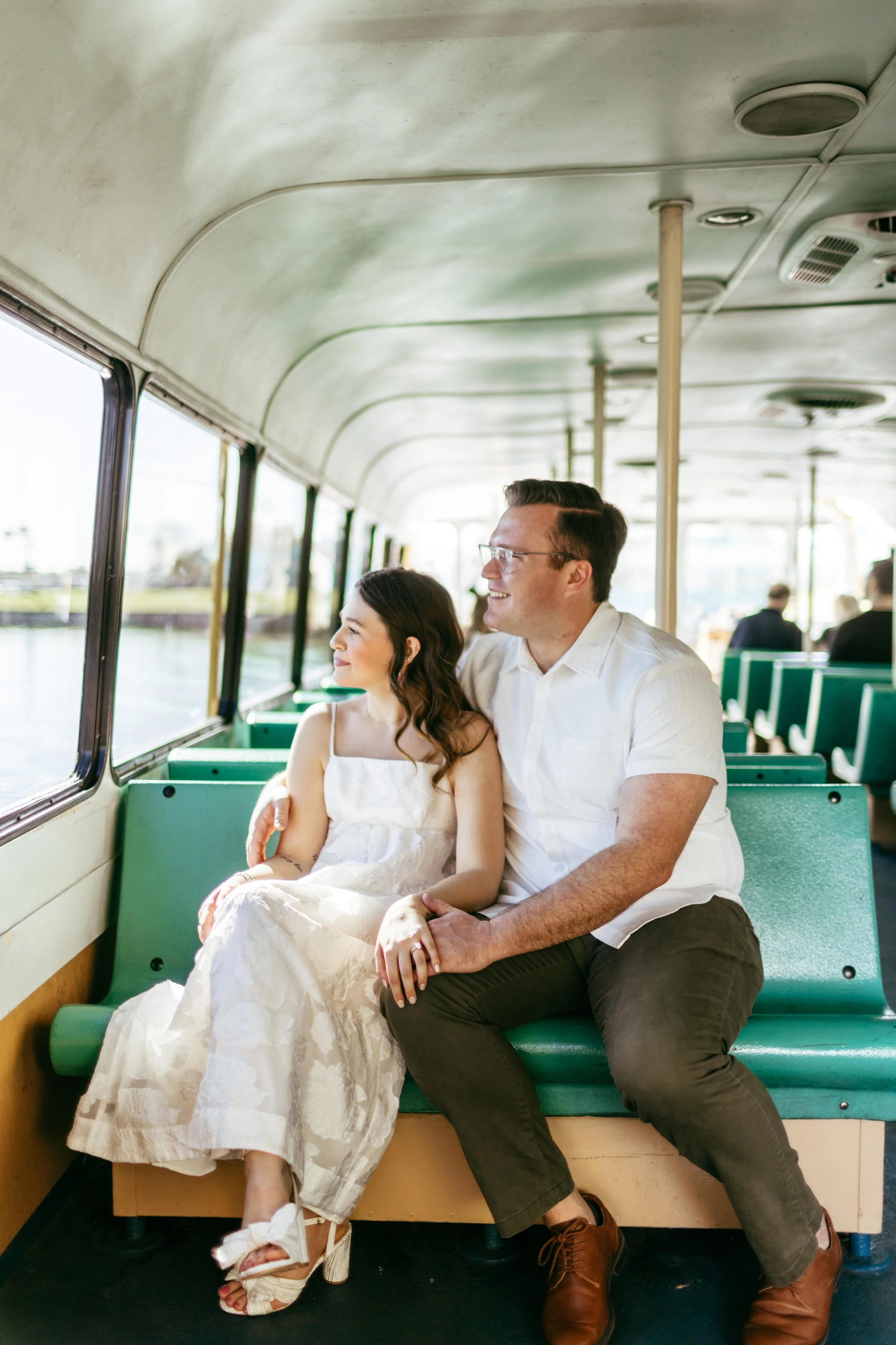A couple sitting on a green bench inside a boat, looking out the window at the water, enjoying a sunny day.