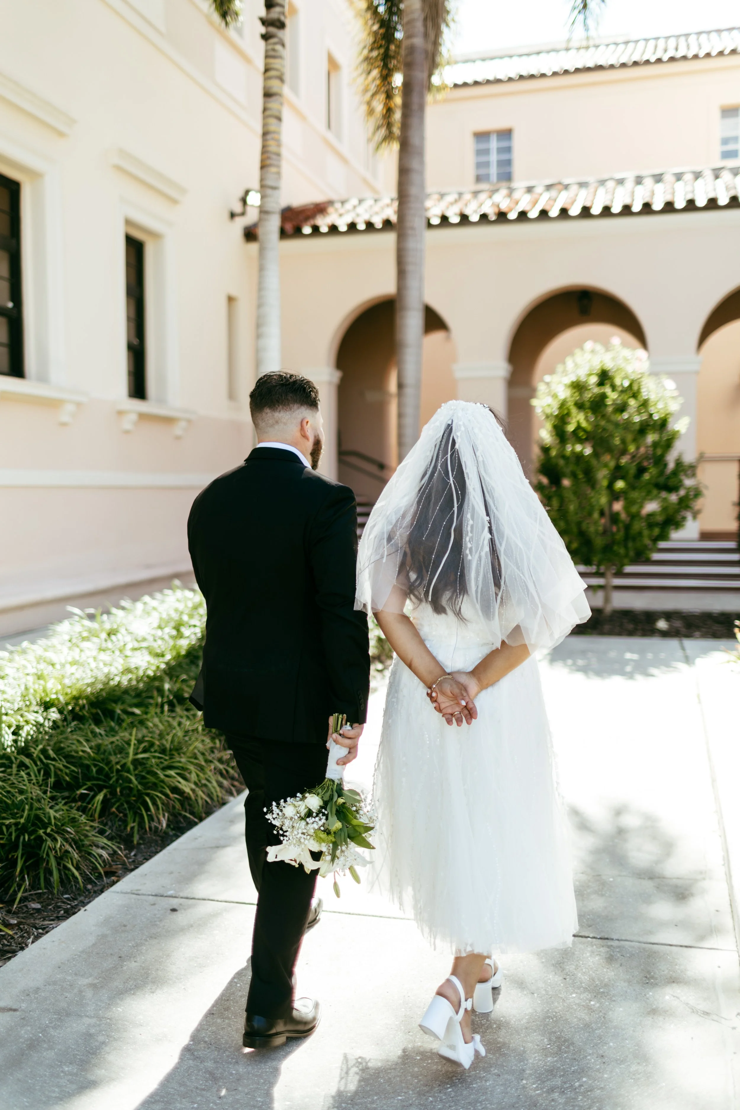 A bride and groom standing together outdoors, with the bride wearing a white wedding dress and veil, and the groom in a black suit holding a bouquet of white flowers.