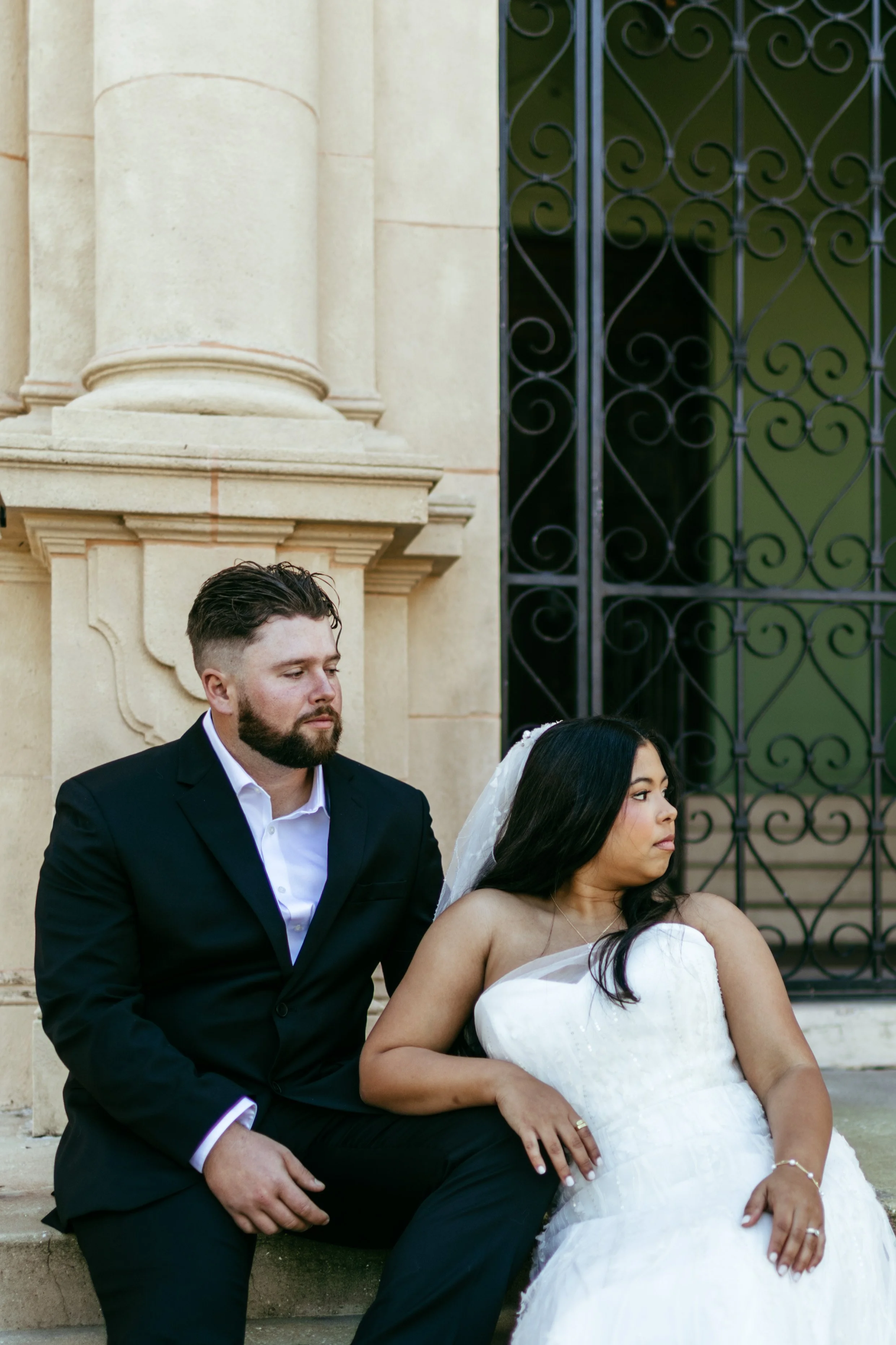 A couple dressed in wedding attire sitting on steps outside a building with stone columns and wrought iron gates.