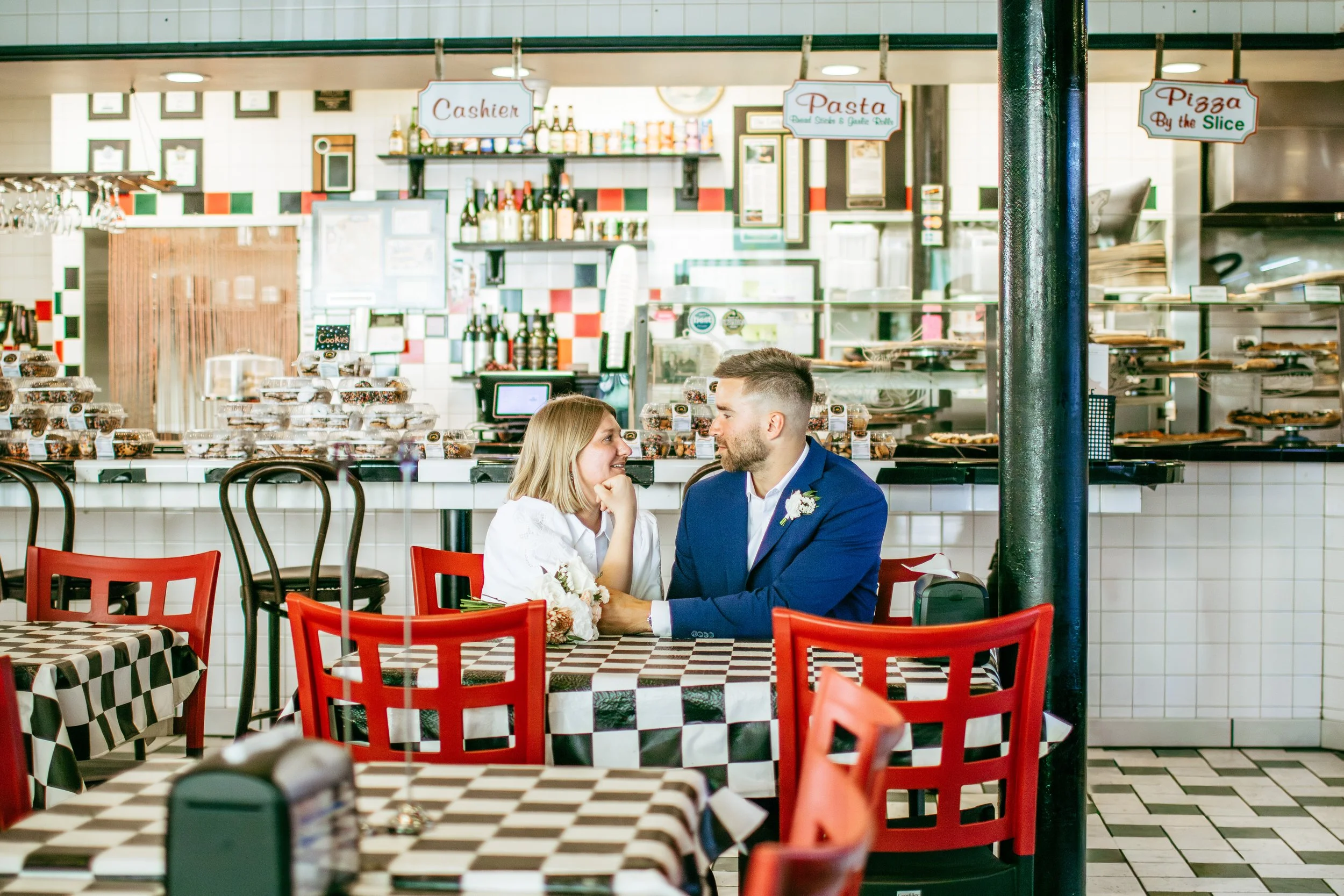 A couple sitting at a checkered table in a restaurant, gazing at each other. The woman is wearing a white dress and the man is wearing a blue suit. The restaurant has a counter in the background with signs for pasta, pizza, and cashier.