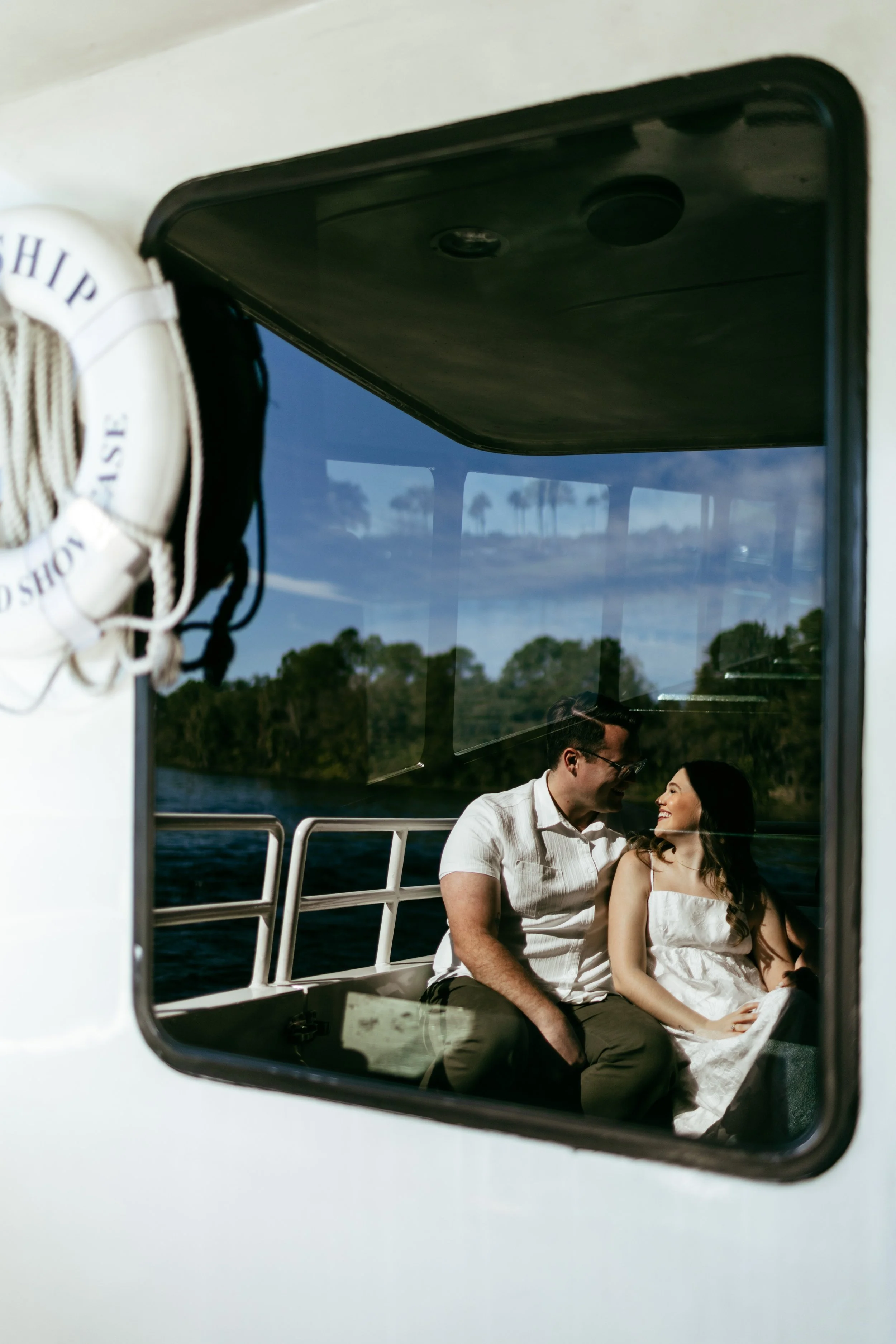 A couple sitting on a boat, looking at each other and smiling, with water and trees visible through the boat window.