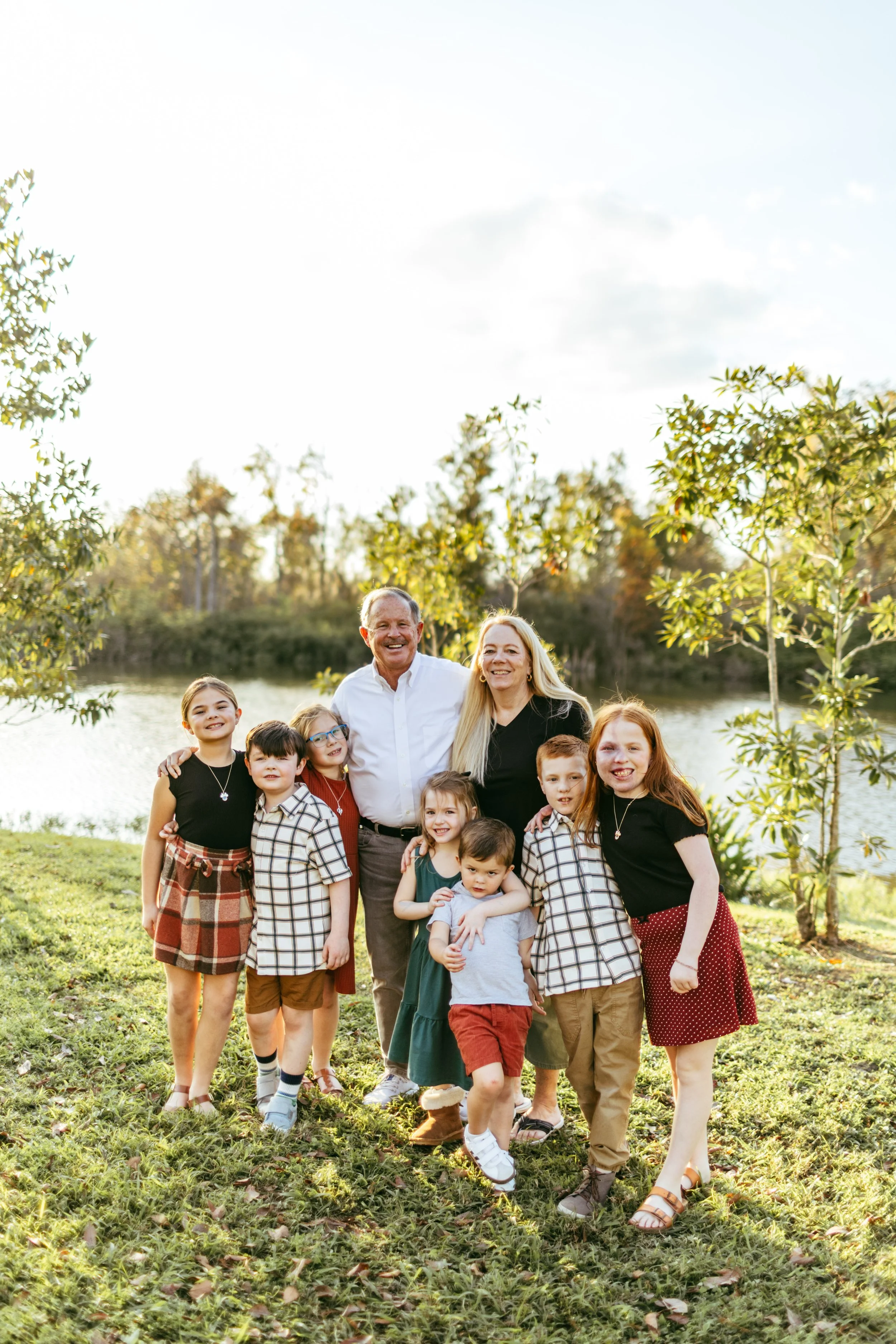 A happy family of nine, including children and adults, posing outdoors by a lake and surrounded by trees during late afternoon or early evening.