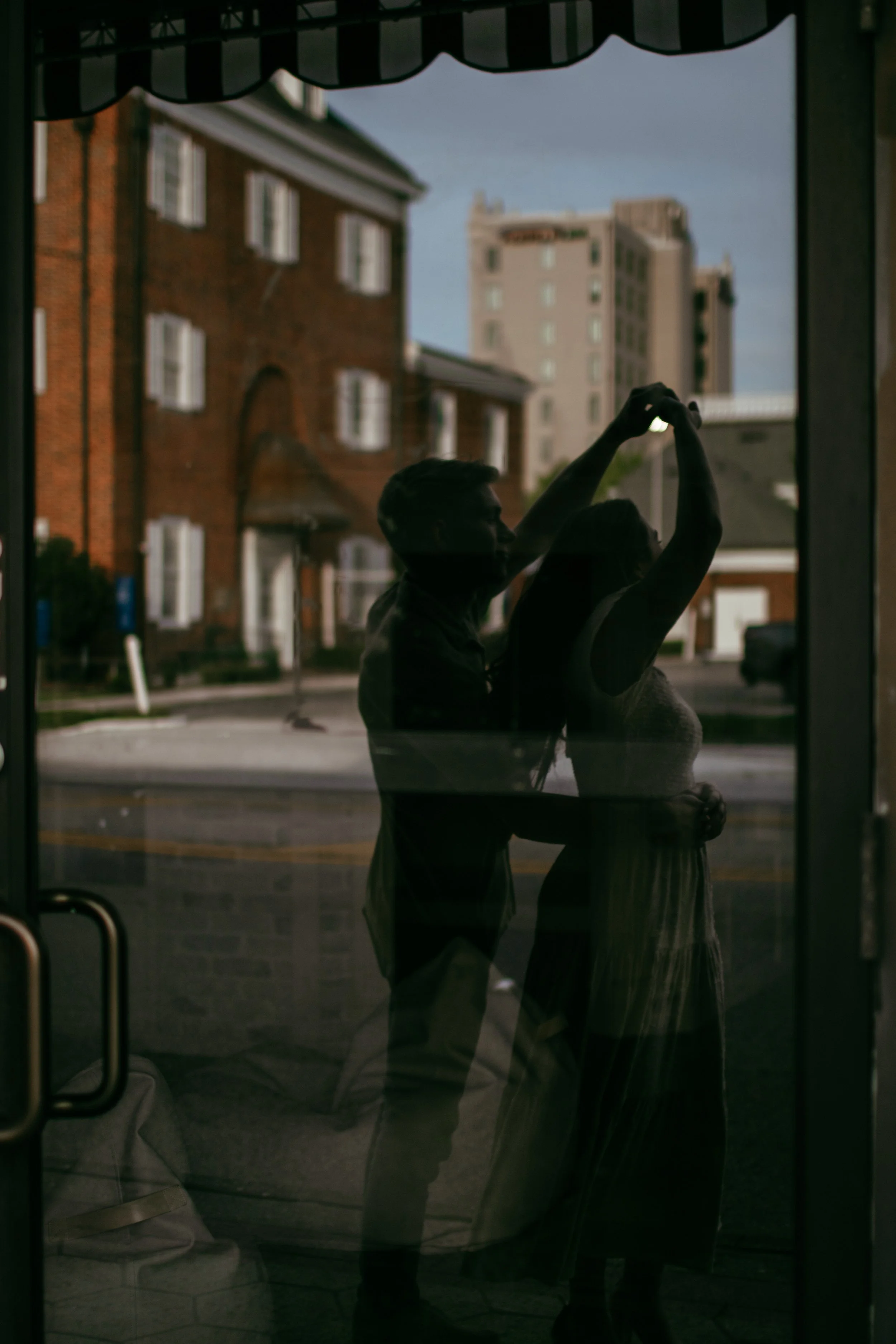 A reflection of a couple dancing, captured through a glass window during daylight, with buildings and a street visible outside.