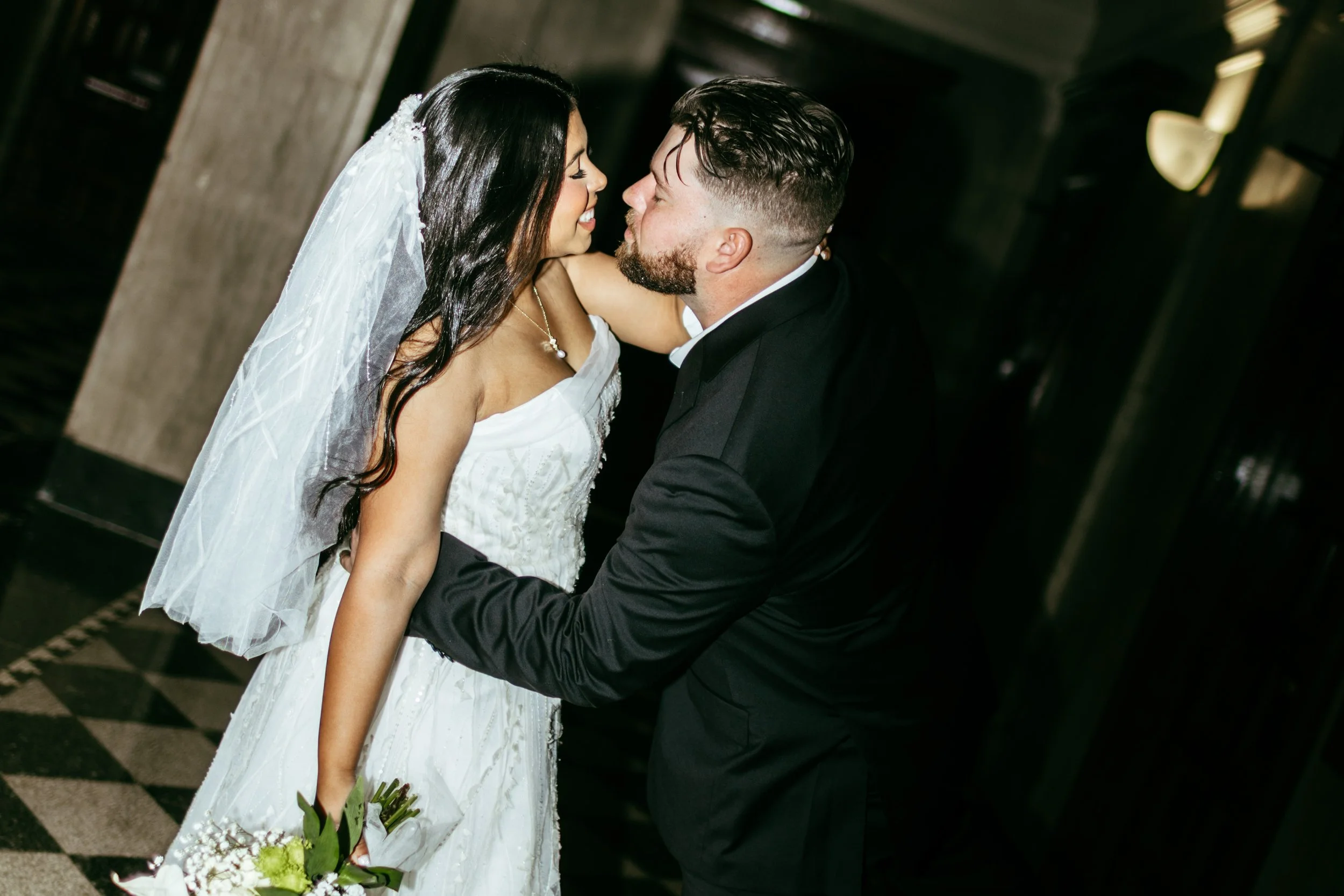A bride and groom sharing a kiss at their wedding, the bride in a white dress and veil, and the groom in a black suit, in a dark indoor setting.