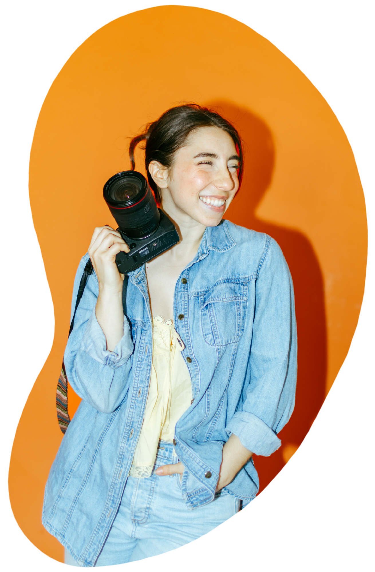 A woman with dark hair wearing a denim jacket, smiling and looking to her right, holding a camera with a strap over her shoulder, standing in front of an orange background with a colorful digital glitch effect.
